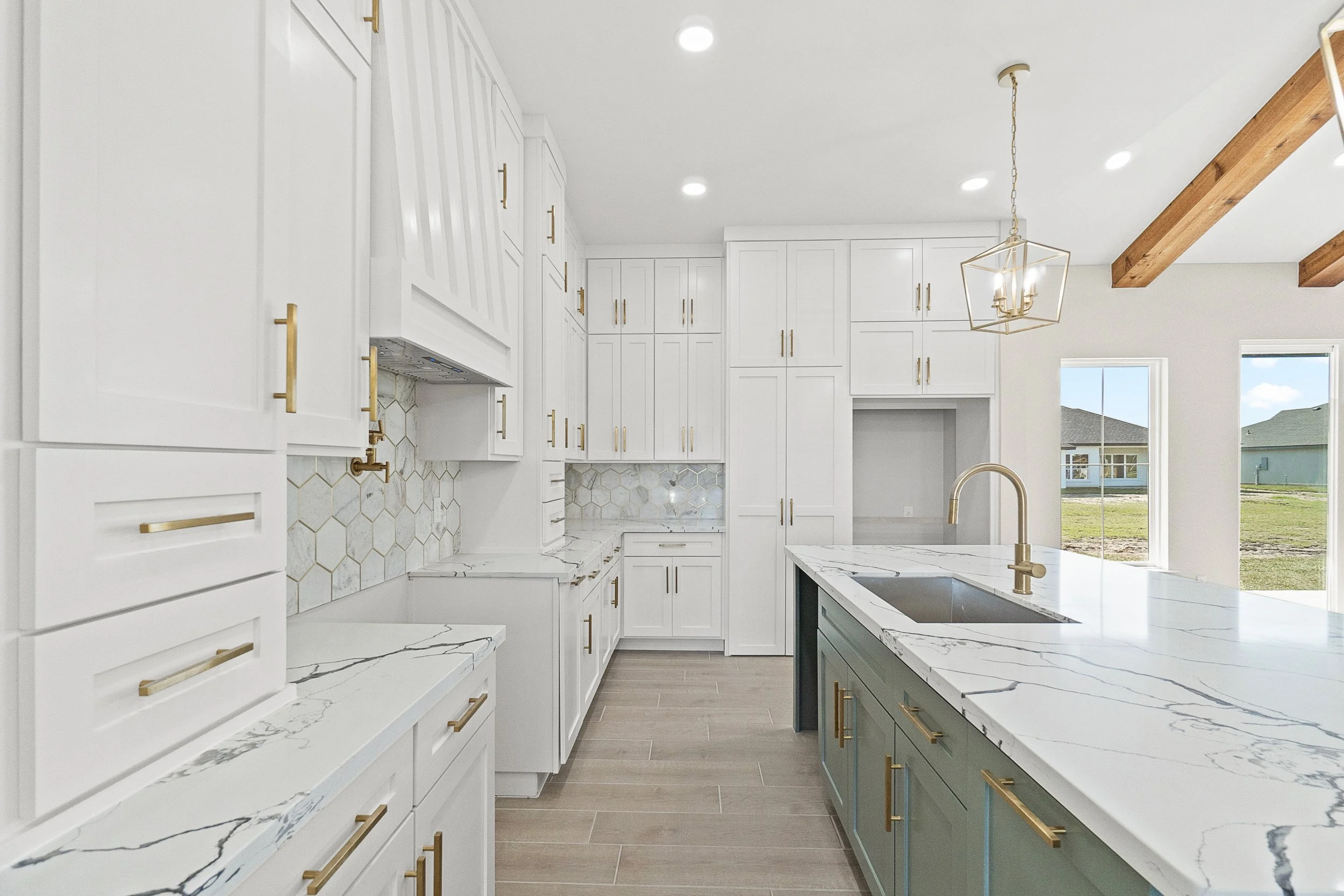 Modern kitchen with white cabinets, gold handles, a large island with a marble countertop, and a gold faucet. There is a hexagonal marble backsplash, and natural light coming through large windows.
