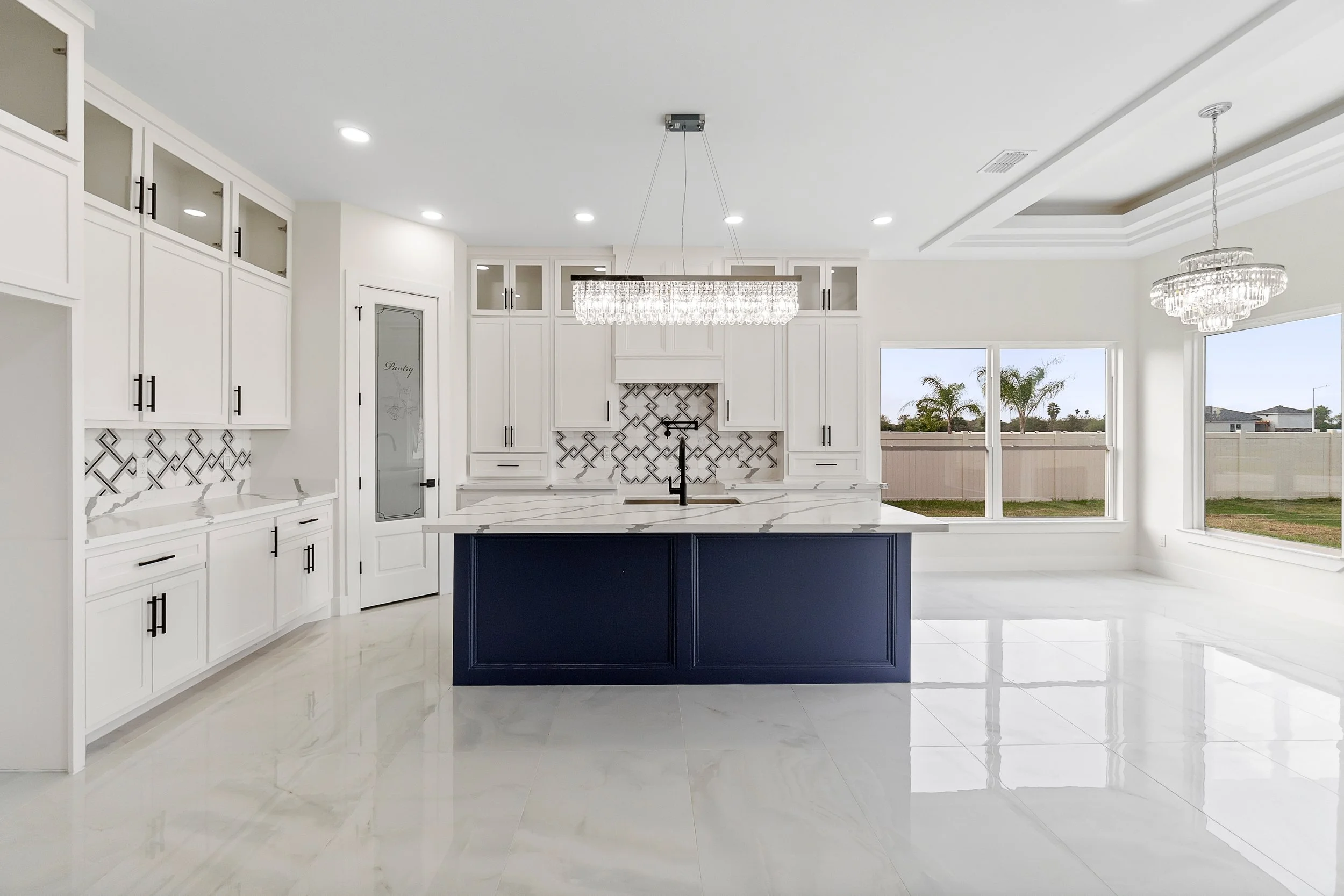 Modern kitchen with white cabinets, a navy blue island, marble countertops, and large windows showing a backyard with palm trees.