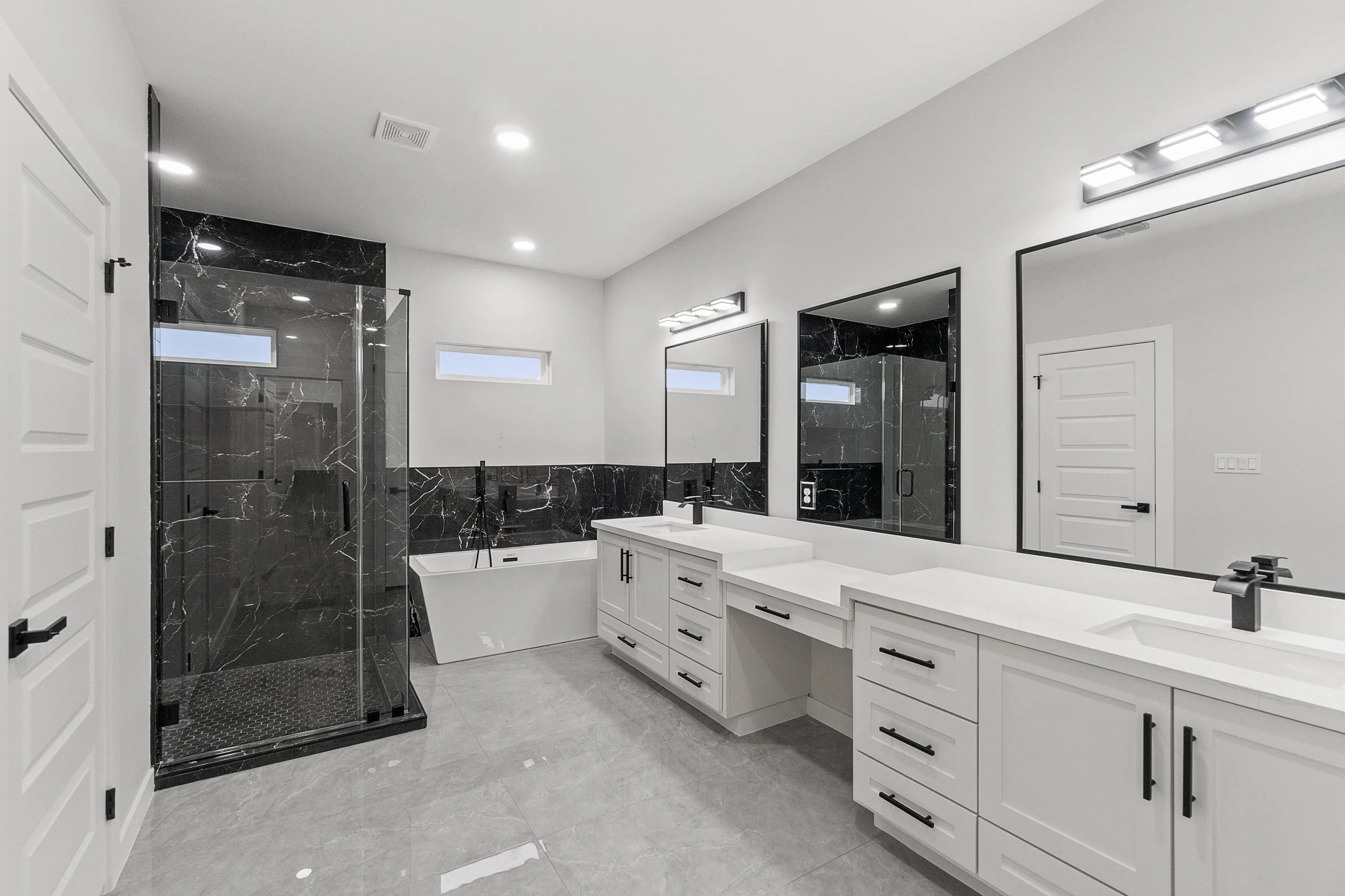 Modern bathroom with large white vanity, dual sinks, black fixtures, and black marble accents. Features a walk-in shower with glass doors, a soaking bathtub, and a white door. Recessed ceiling lights and horizontal windows provide illumination and na