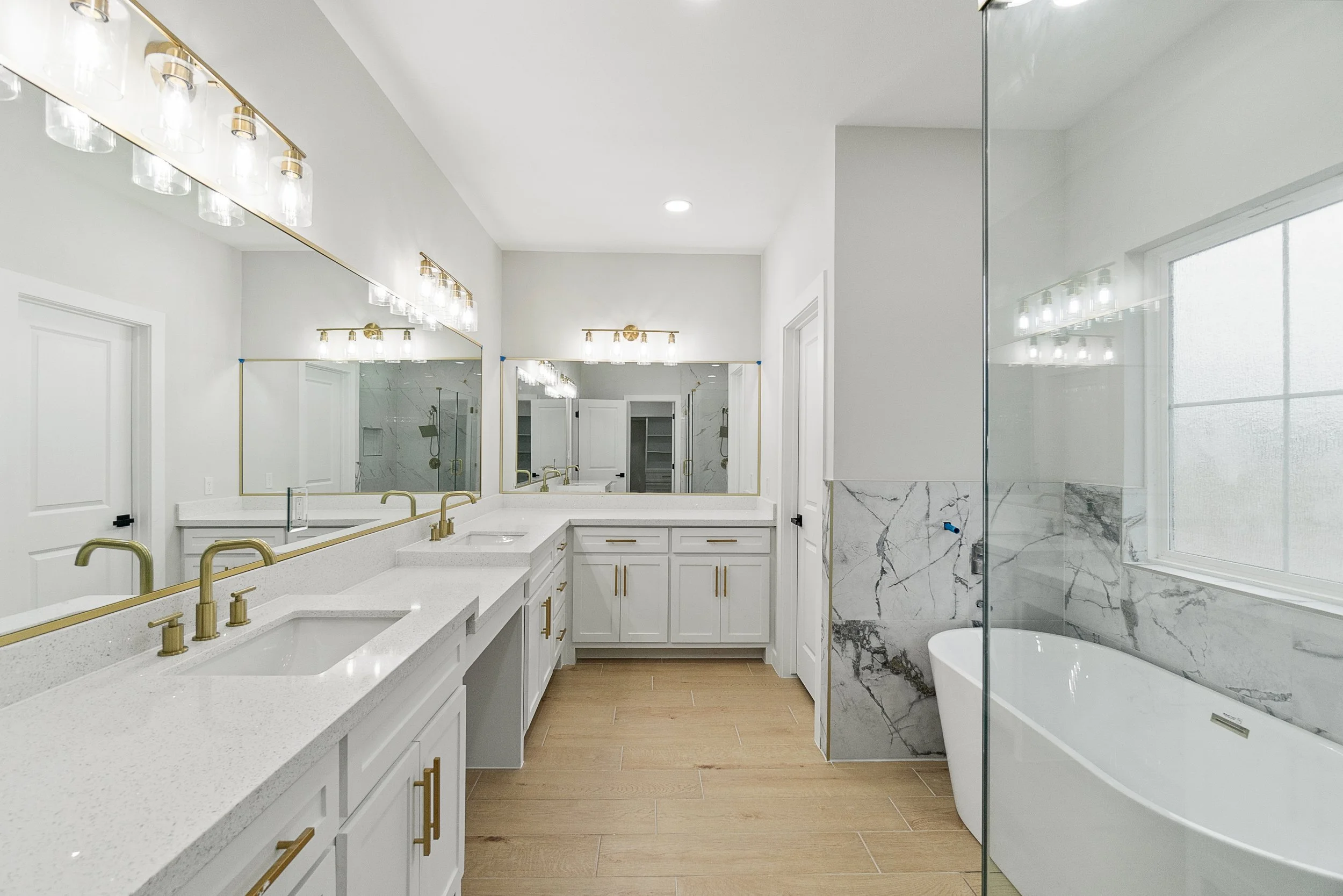 Modern bathroom with white cabinets, gold fixtures, large mirrors, and a bathtub next to a window