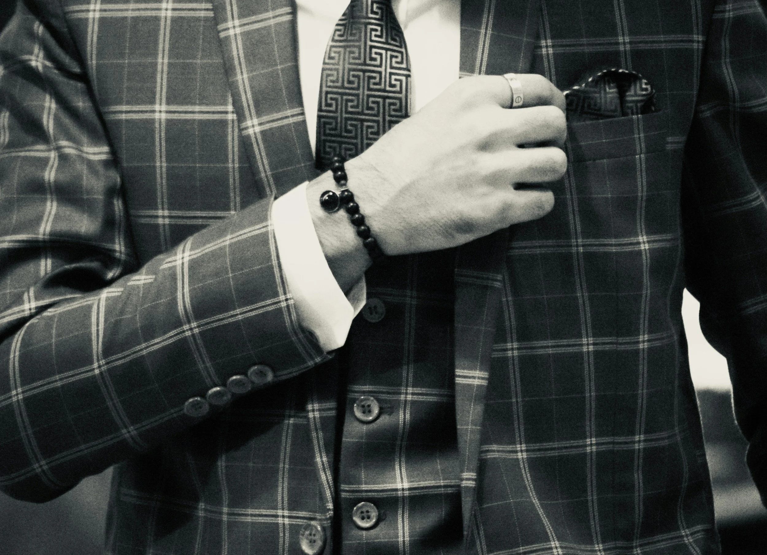 Close-up of a man in a checkered suit adjusting his shirt collar, wearing a patterned tie, with a bracelet on his wrist and a ring on his finger.