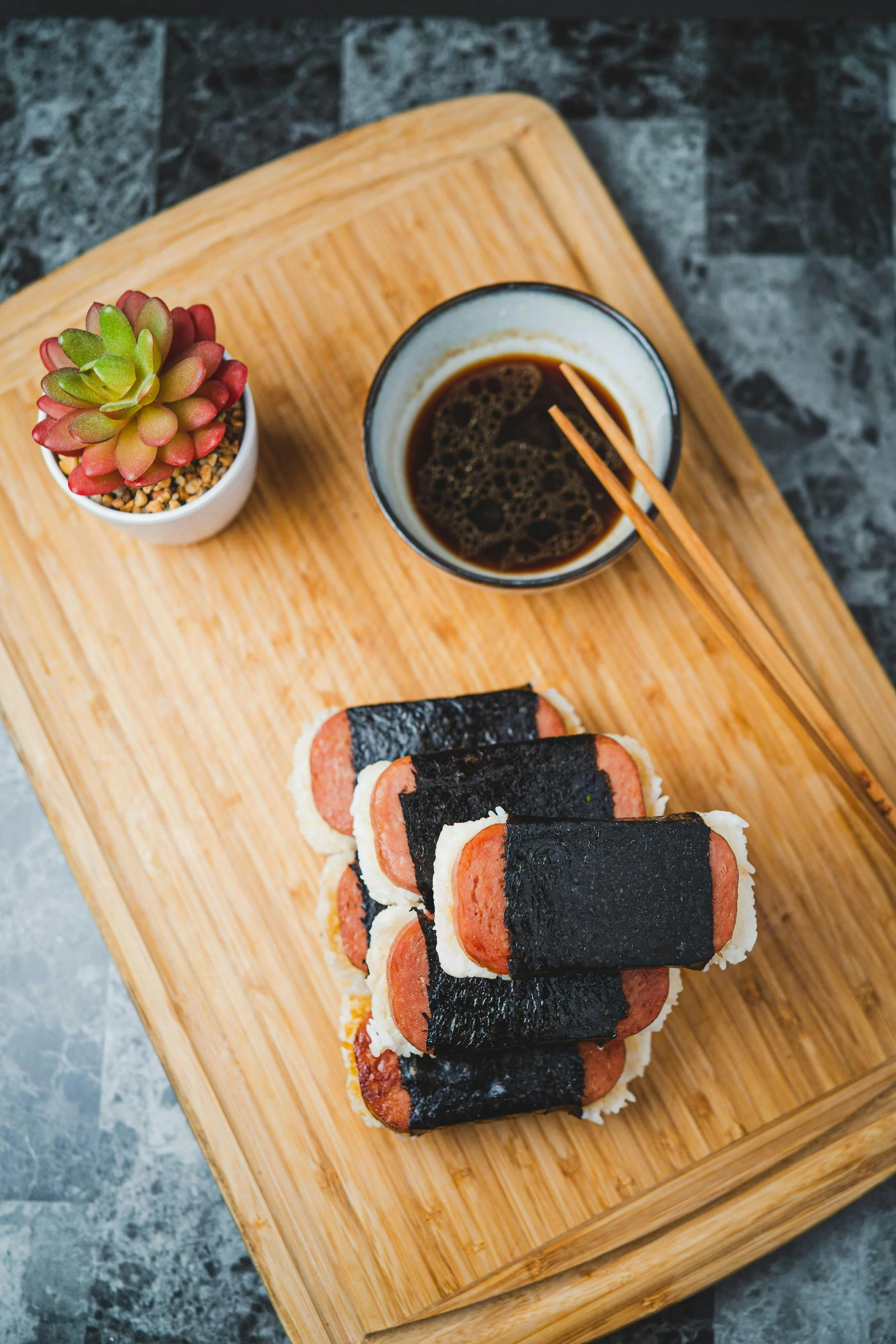 A bamboo tray holds a cup of black coffee with wooden chopsticks, several sushi rolls wrapped in seaweed with sausage and rice, and a small potted succulent plant.