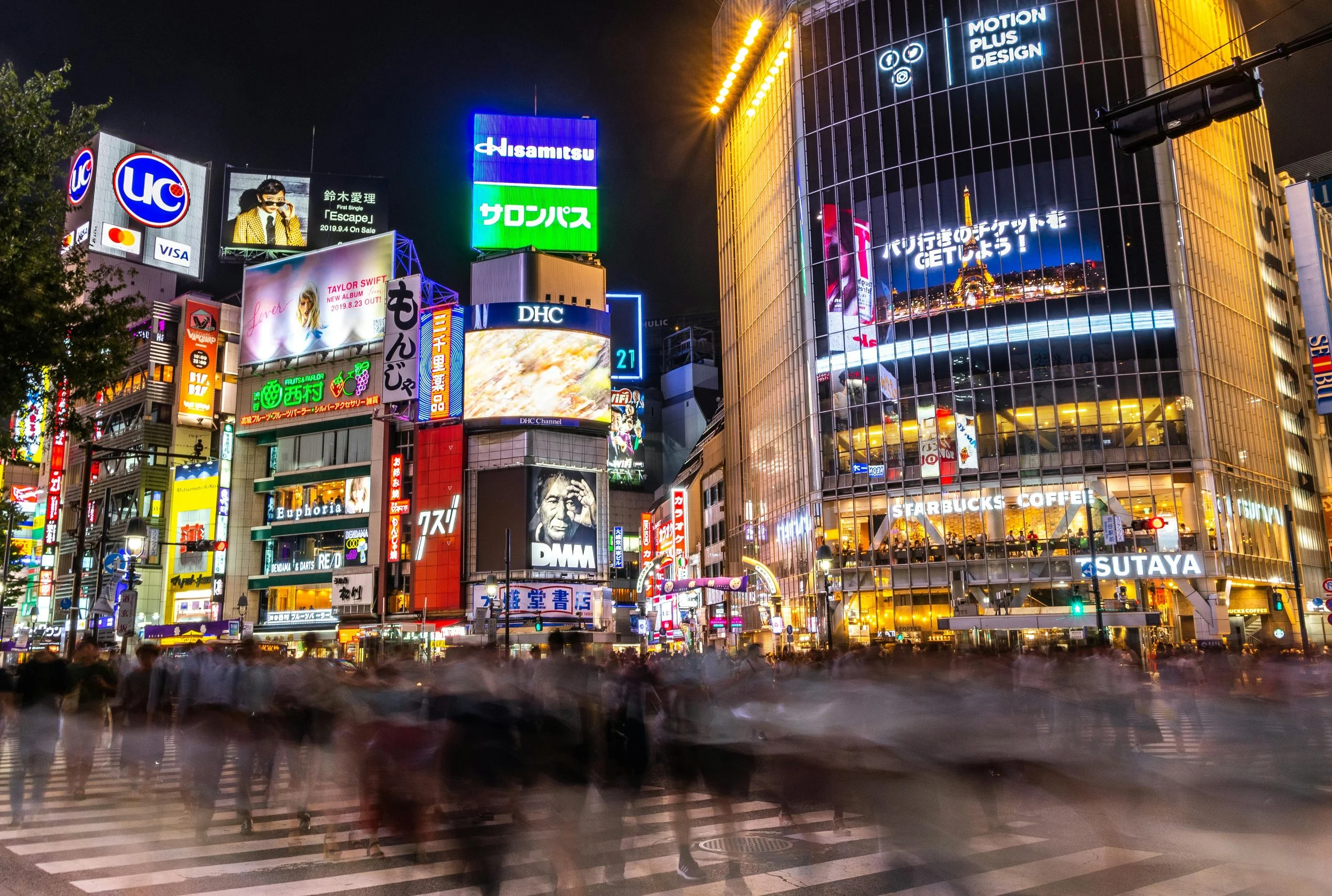 Nighttime cityscape of a busy intersection in Tokyo, Japan, with illuminated billboards, neon signs, and a glass-fronted Starbucks coffee shop, capturing the vibrant nightlife and crowd movement.