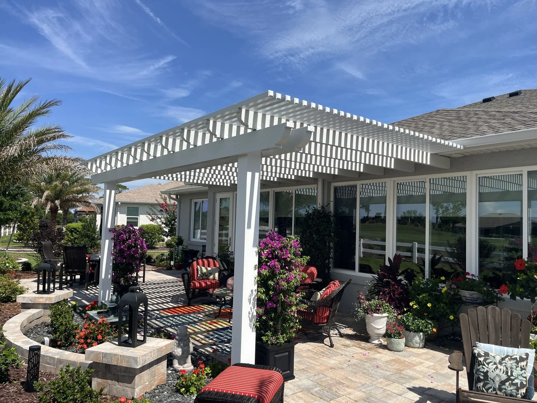 A backyard patio with outdoor furniture, potted plants, and purple flowers under a white pergola, with a house and a lawn in the background.