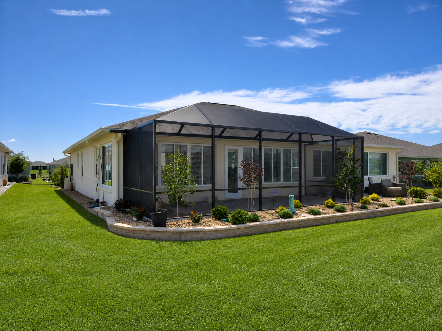 Backyard of a house with a screened-in patio, green lawn, and garden bed with plants and trees under a partly cloudy blue sky.