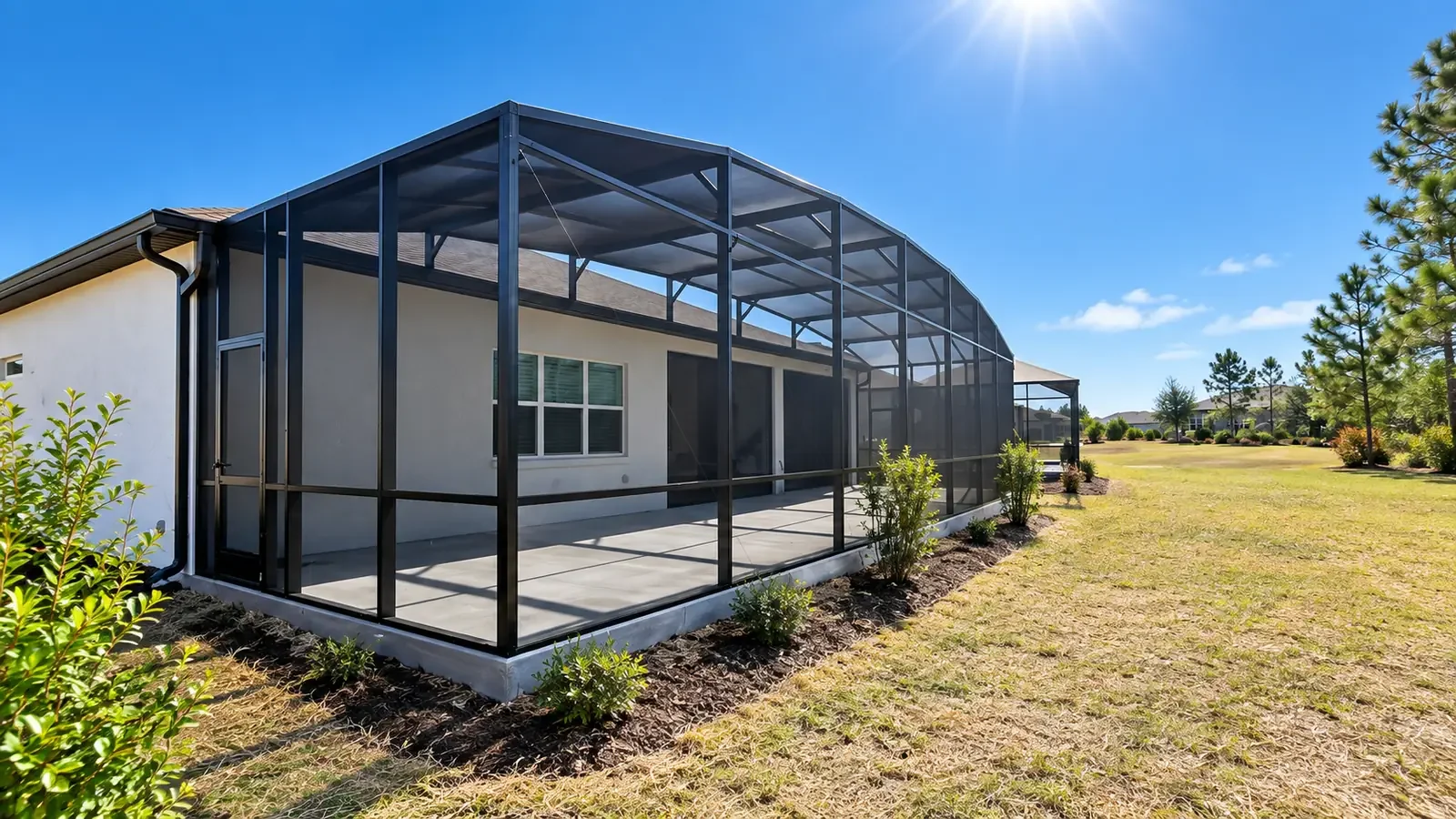 Backyard with screen cage, outdoor furniture, and a house in the background with a clear blue sky and some clouds.