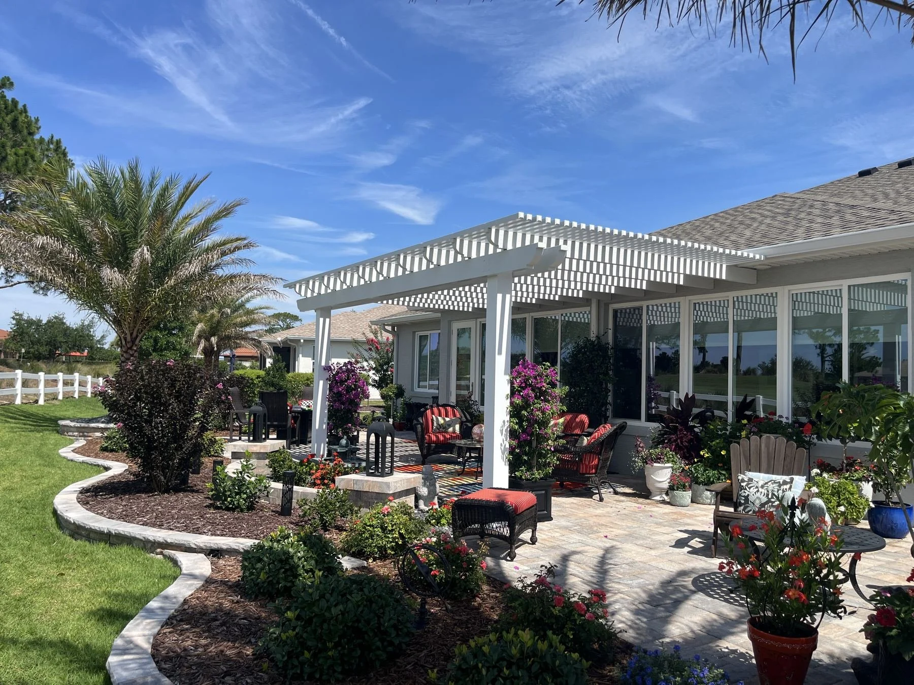 A backyard patio with outdoor furniture, potted plants, and purple flowers under a white pergola, with a house and a lawn in the background.