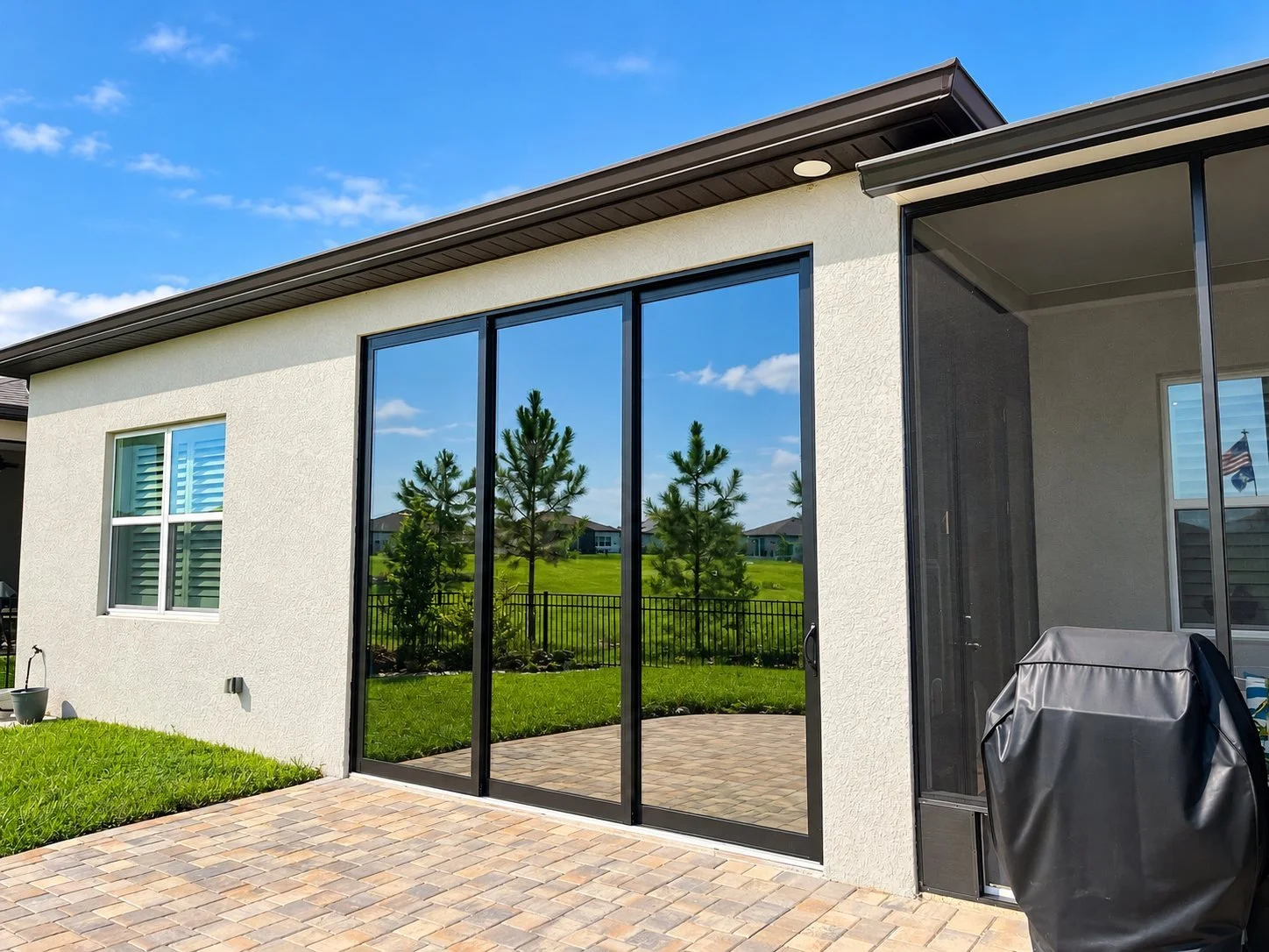 Backyard backyard with a sliding glass door, small window with shutters, barbecue grill covered with a black cover, brick paver patio, lawn with trees, and neighboring houses
