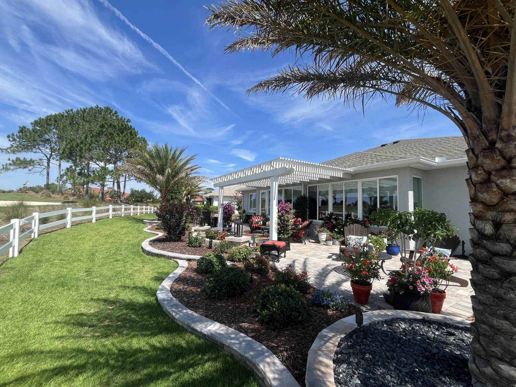 A backyard patio with a gazebo, outdoor furniture, potted plants, and a landscaped garden, with trees and a blue sky with wispy clouds in the background.