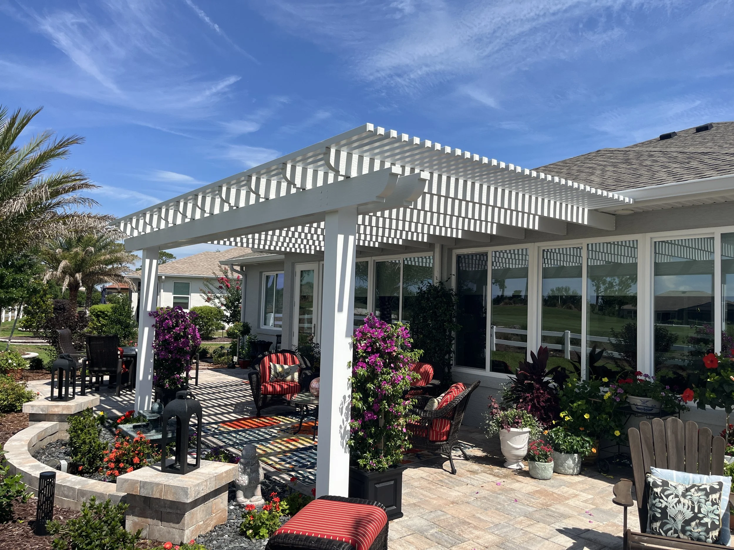 A backyard patio with outdoor furniture, potted plants, and purple flowers under a white pergola, with a house and a lawn in the background.