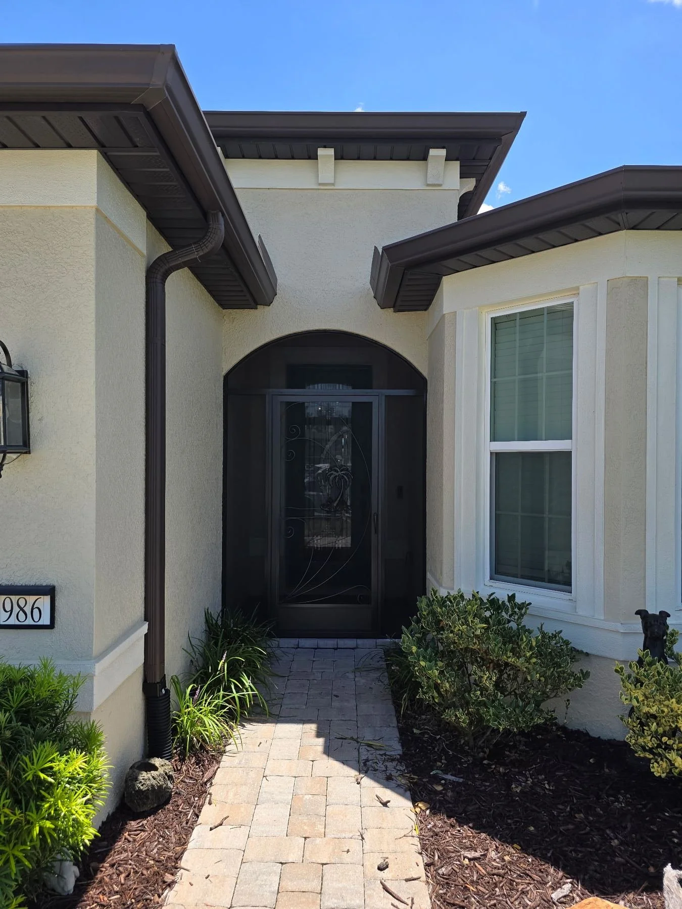 Front entrance of a house with a brick pathway leading to a black screen door, surrounded by green shrubbery and plants, with a window on the right side and a house number 986 on the left.