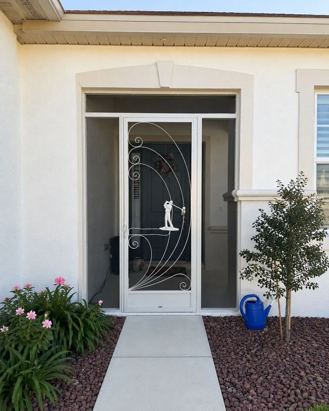 Front entrance of a house with a decorative screen door featuring a sculpture of a person fishing, set against a dark interior door. There are pink flowers and green plants on either side of the walkway, with a blue watering can next to a small tree 