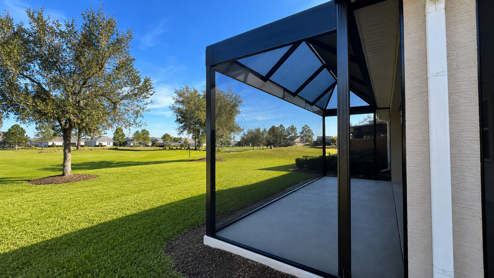 A screened porch with black framing attached to a beige house, overlooking a green lawn with trees and a neighborhood in the distance, under a blue sky.