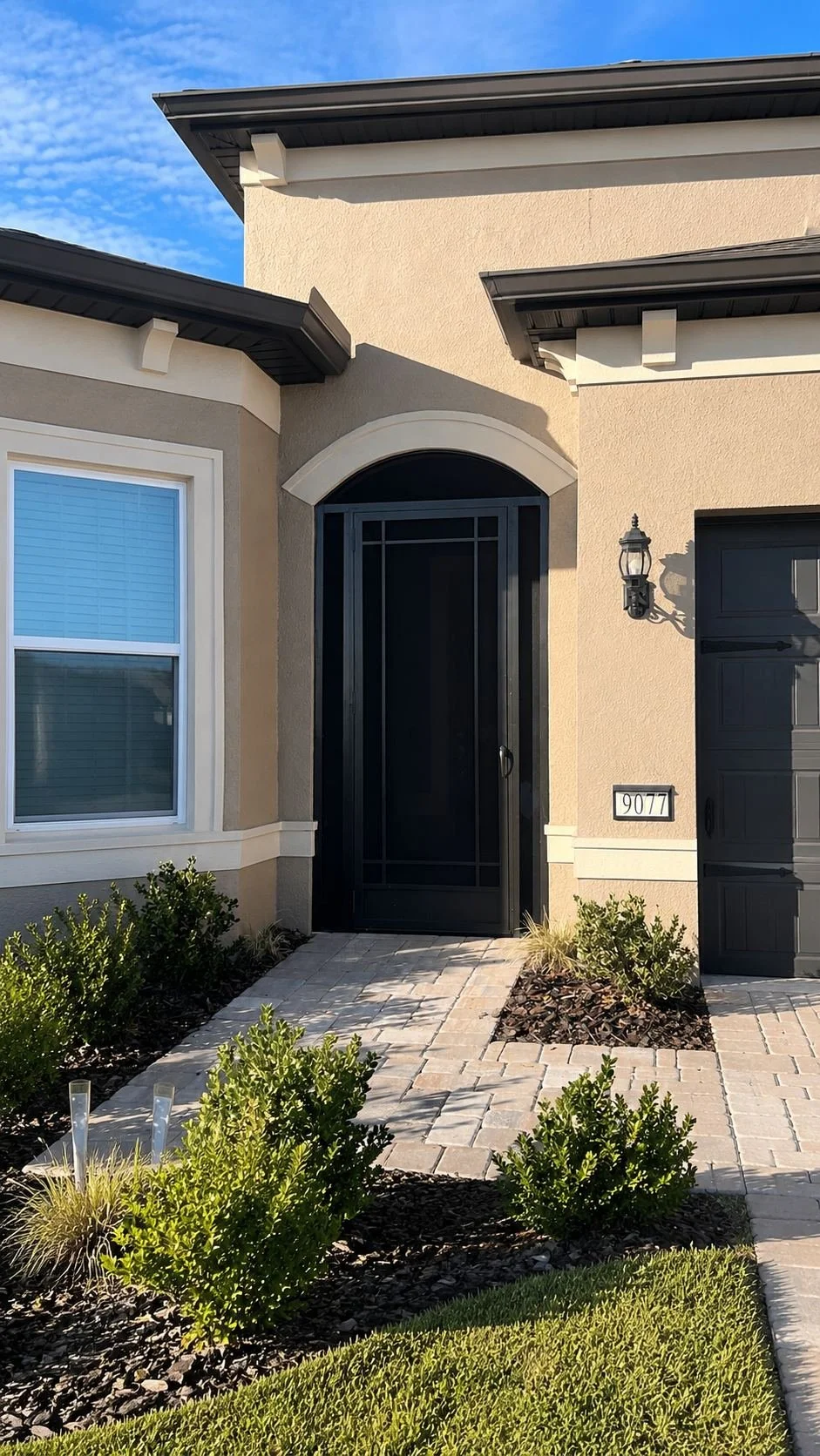 Modern house entrance with black door and security screen, landscaped garden with shrubs and brick pathway, house number 9077, exterior wall lantern, clear blue sky.
