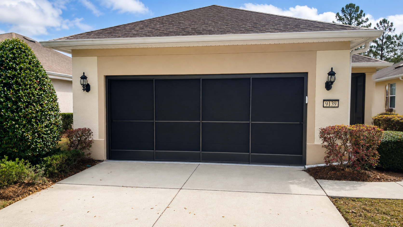 Front view of a house with sliding garage screen doors, beige exterior walls, and outdoor wall-mounted lantern lights on each side of the door. Surrounding shrubbery and a concrete driveway leading up to the garage.