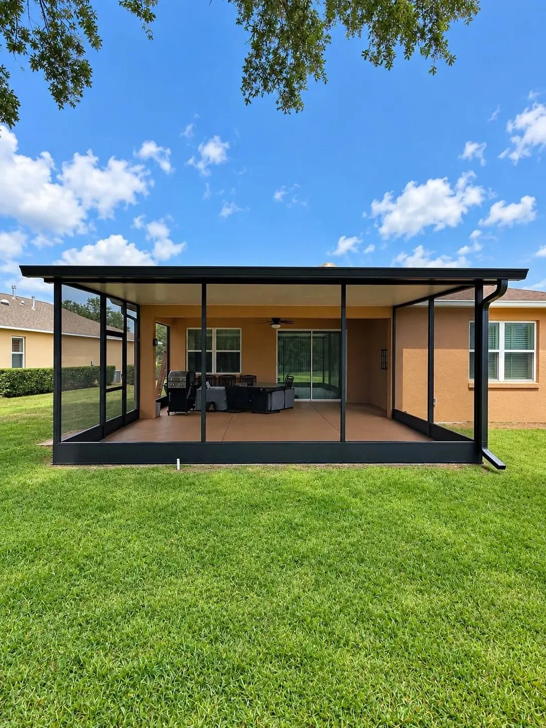 A screened porch with outdoor furniture, ceiling fan, and sliding glass door, attached to a beige house with a green lawn and a blue sky with scattered clouds.