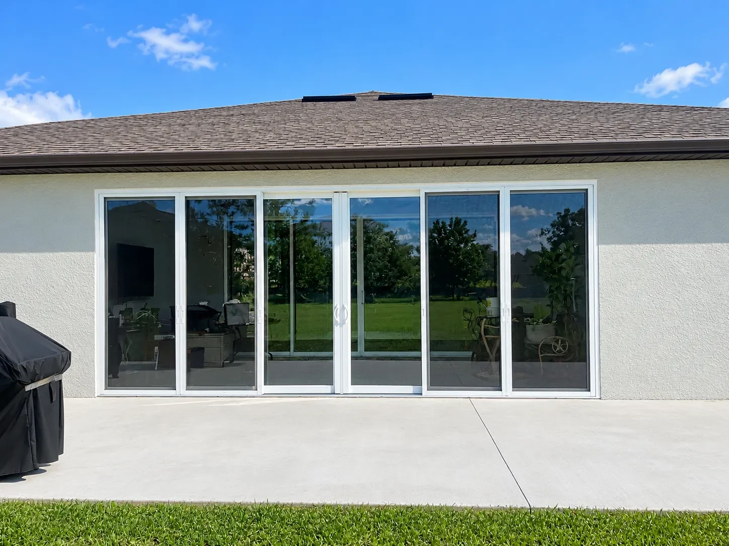 Back yard view of a house with large sliding glass doors, a concrete patio, a covered barbecue grill on the left, green lawn, and trees in the background under a blue sky with some clouds.