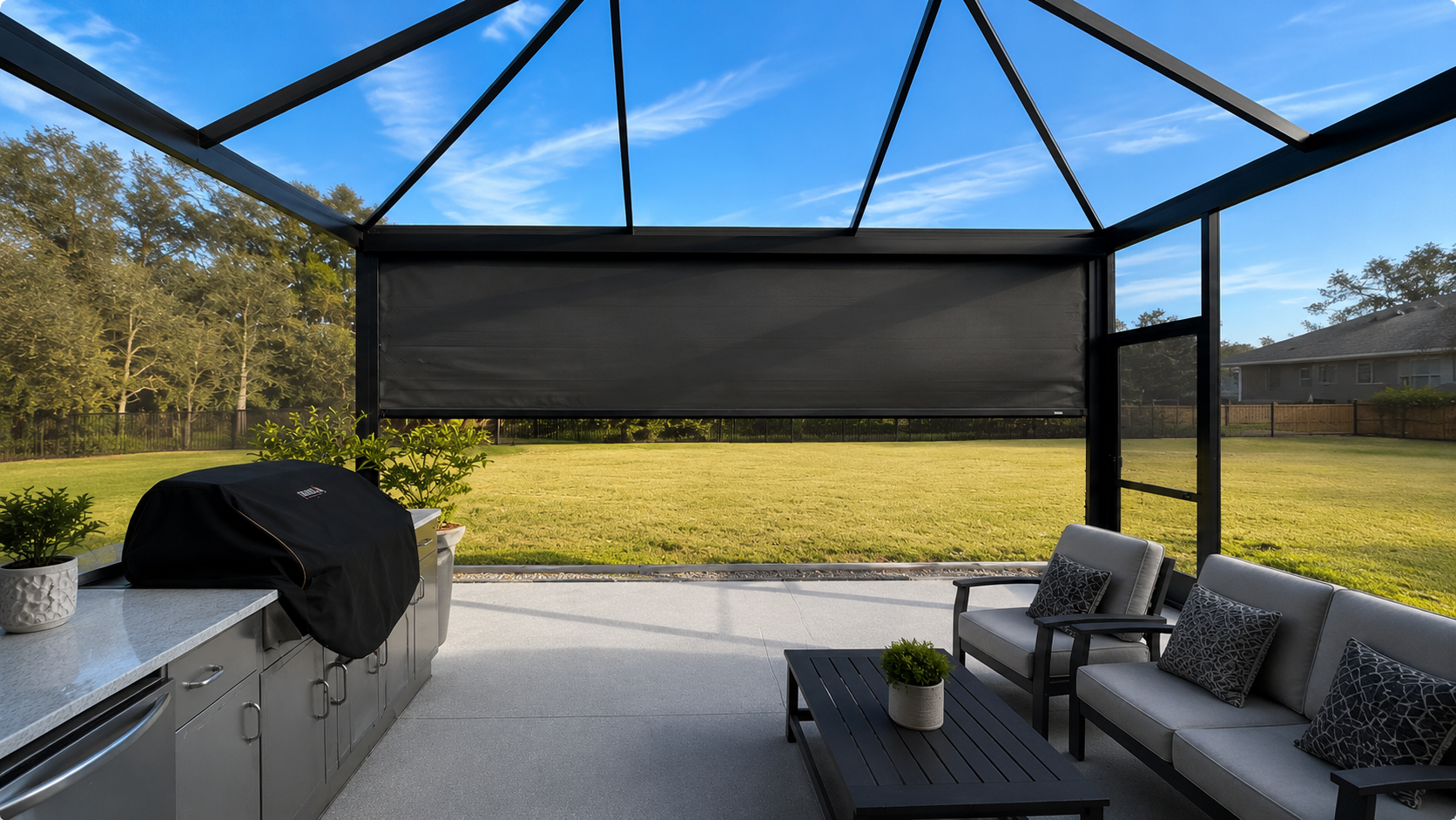 Outdoor patio with seating and a grill a screen cage with a motorized screen unit on the inside, overlooking a grassy backyard and neighboring houses.