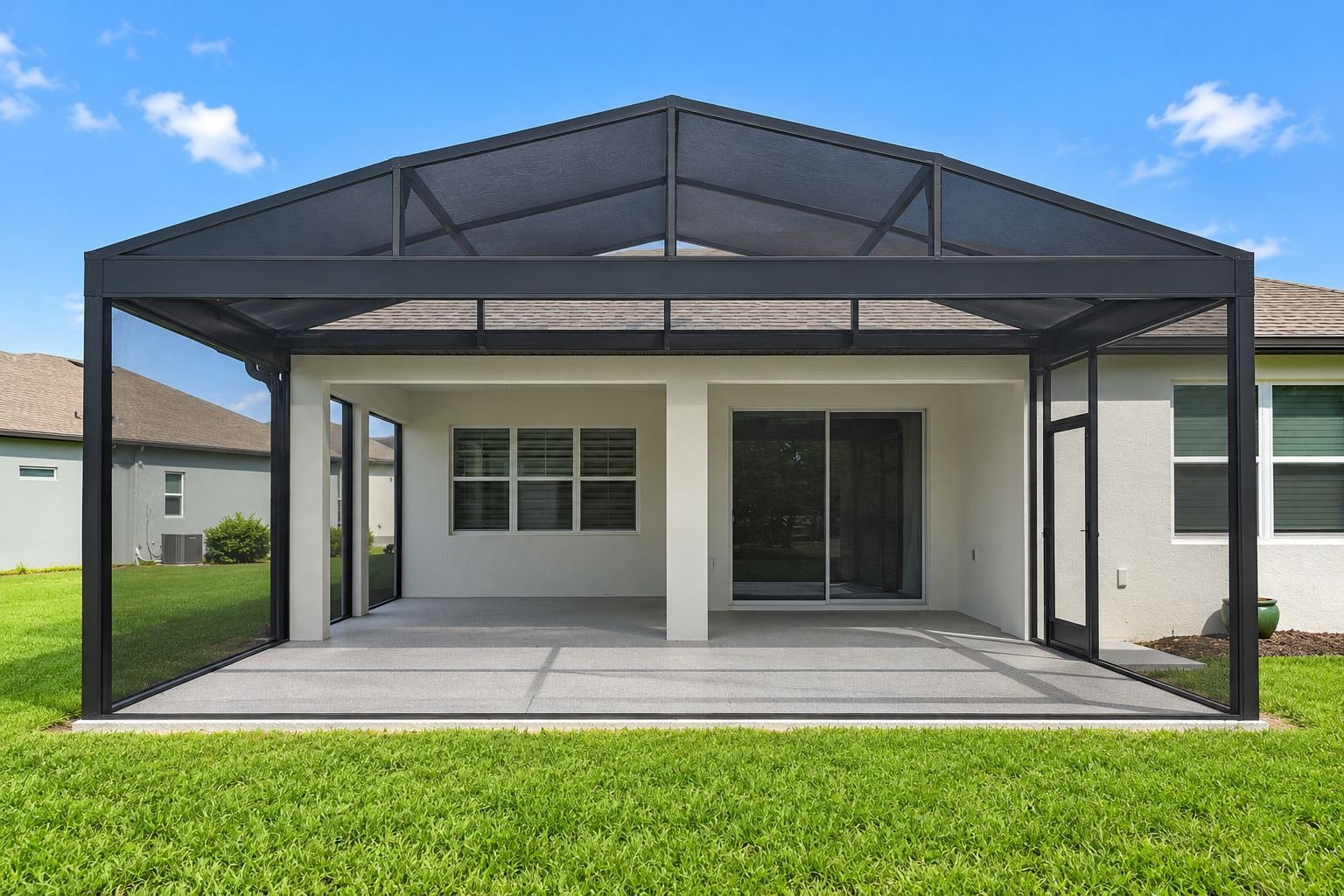 A screened-in porch with a black metal frame attached to a white house, overlooking a green lawn and a neighboring house under a blue sky with few clouds.