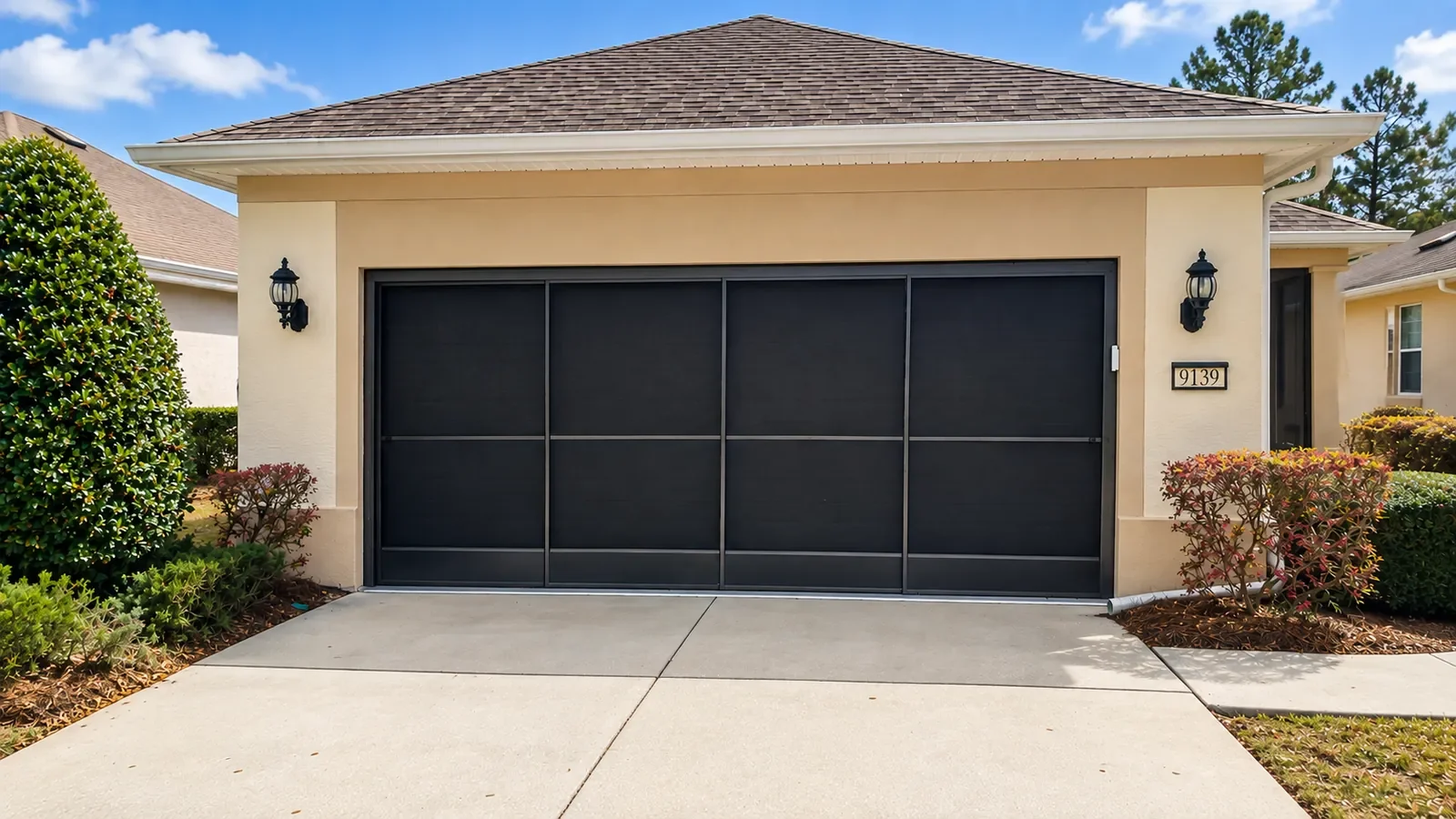 Front view of a house with sliding garage screen doors, beige exterior walls, and outdoor wall-mounted lantern lights on each side of the door. Surrounding shrubbery and a concrete driveway leading up to the garage.
