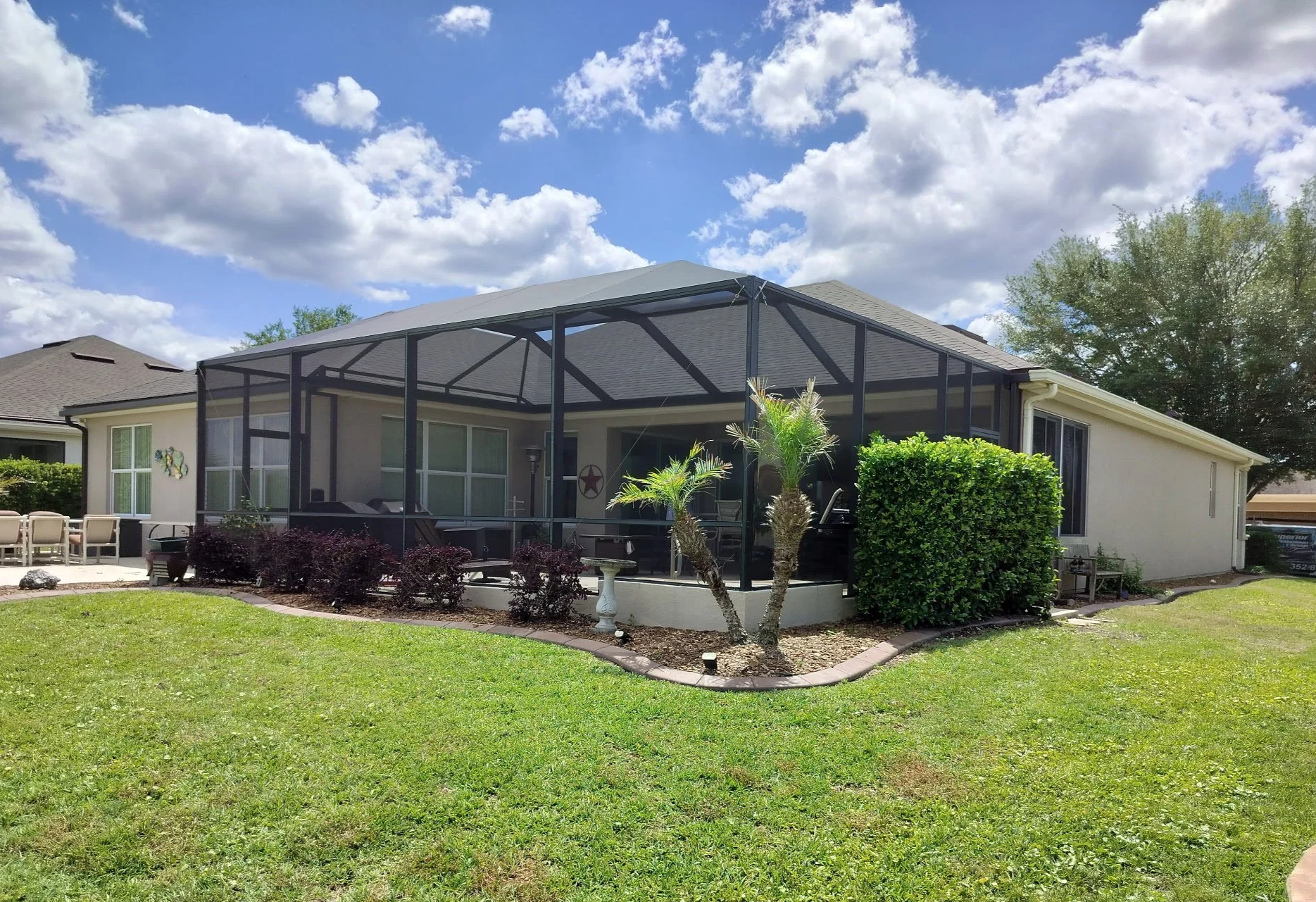 Backyard with a screened-in porch, outdoor seating area, small trees, bushes, and a well-maintained lawn under a partly cloudy sky.