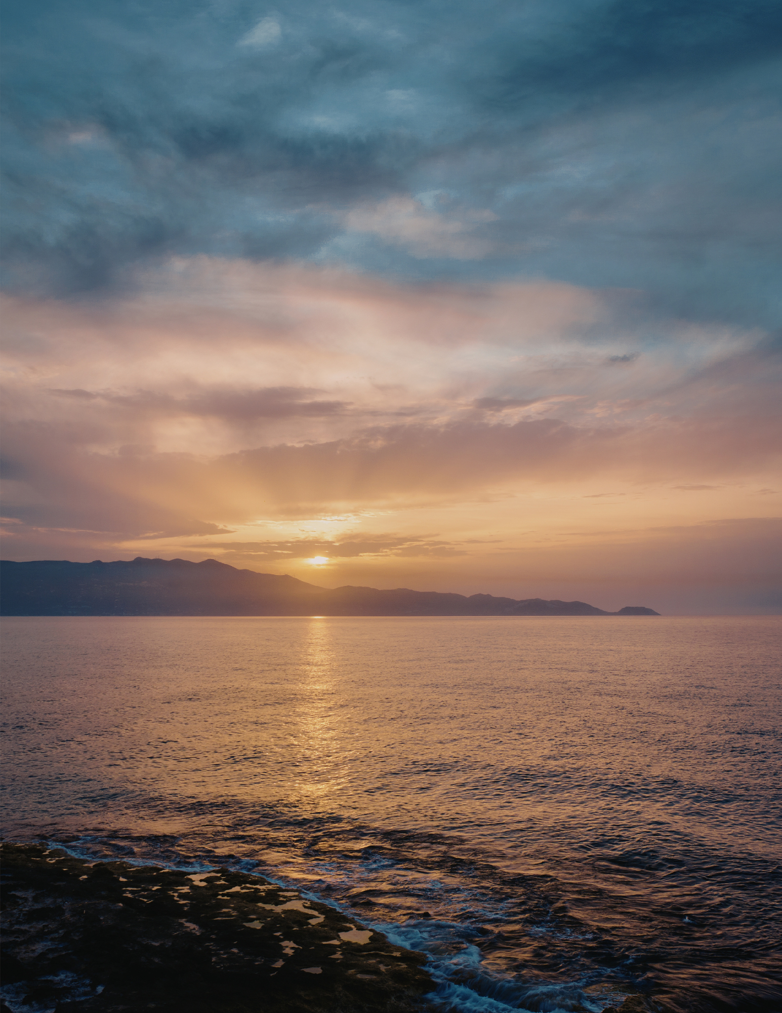 Sunset over the ocean with mountains in the distance and colorful clouds in the sky.