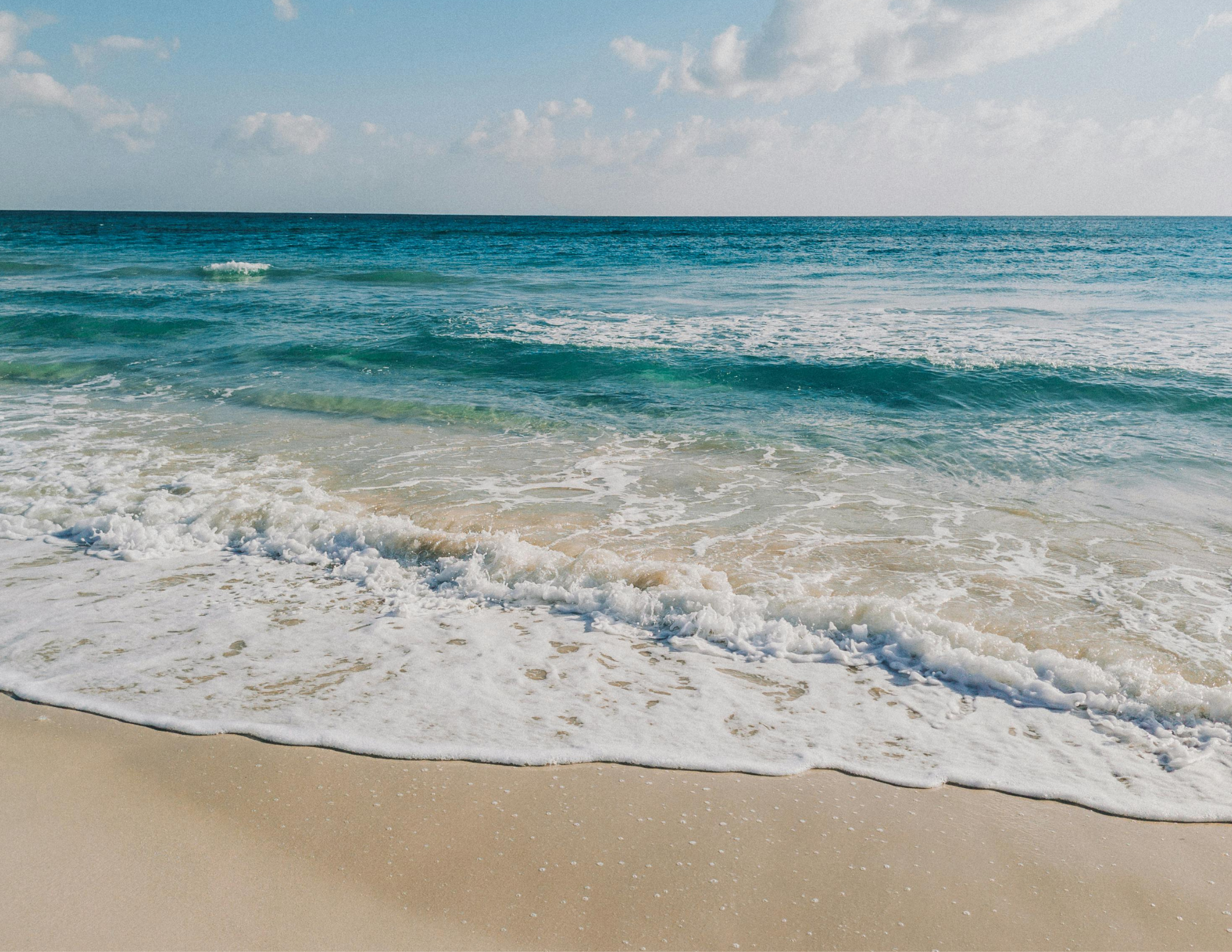Sunny beach with gentle waves lapping sandy shore, clear blue sky with a few clouds.