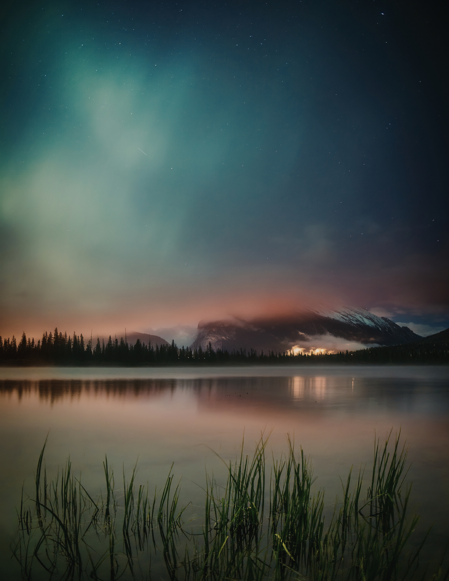 Nighttime landscape photo of a calm lake with reflections, surrounded by pine trees, mountains, and a colorful sky with stars and aurora borealis.
