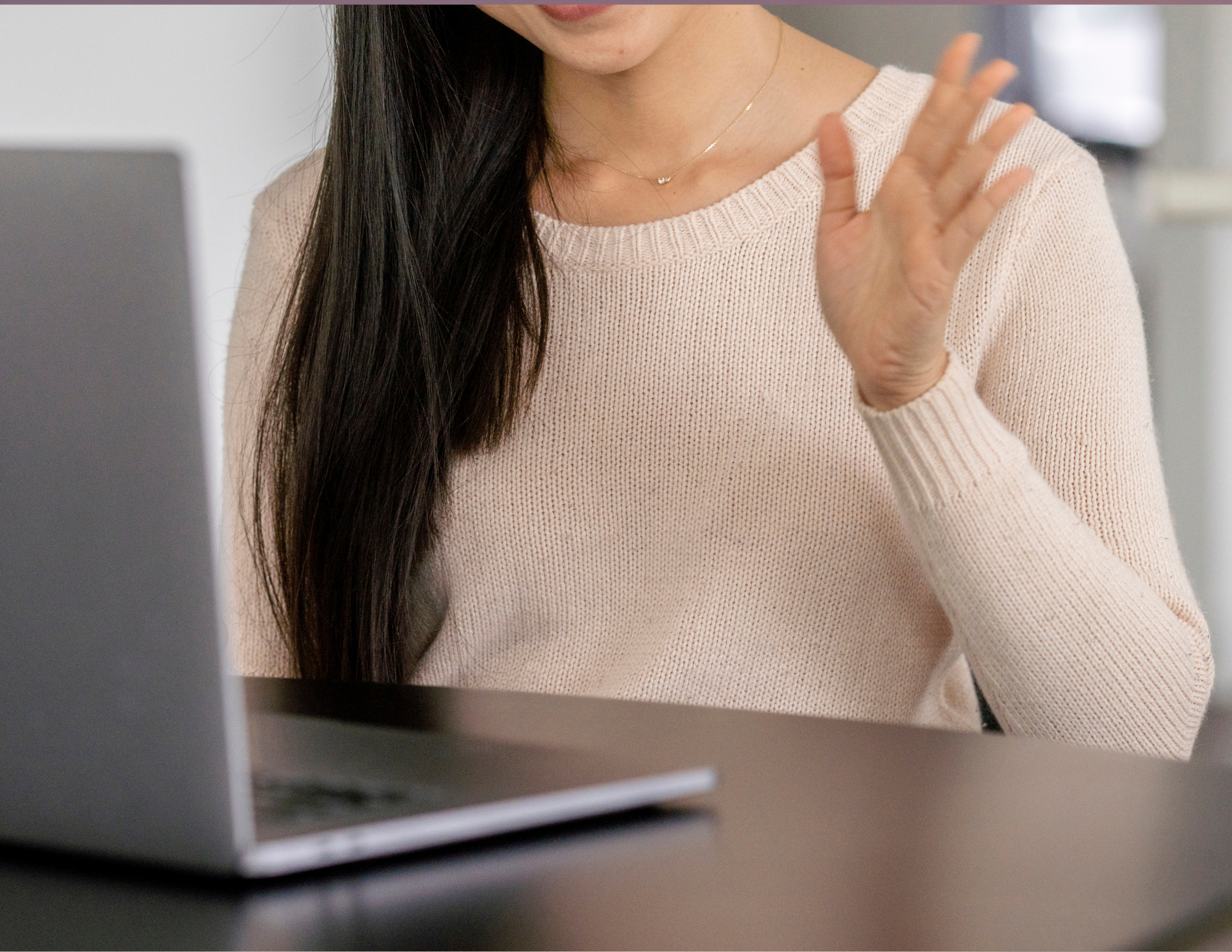 A woman with long dark hair wearing a cream sweater, sitting at a desk with a laptop, holding up her hand in a waving gesture.
