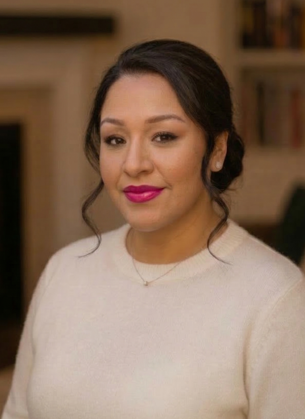Portrait of a woman with dark hair styled in a low bun, wearing a cream sweater, pink lipstick, and small earrings, standing indoors with a bookshelf in the background.