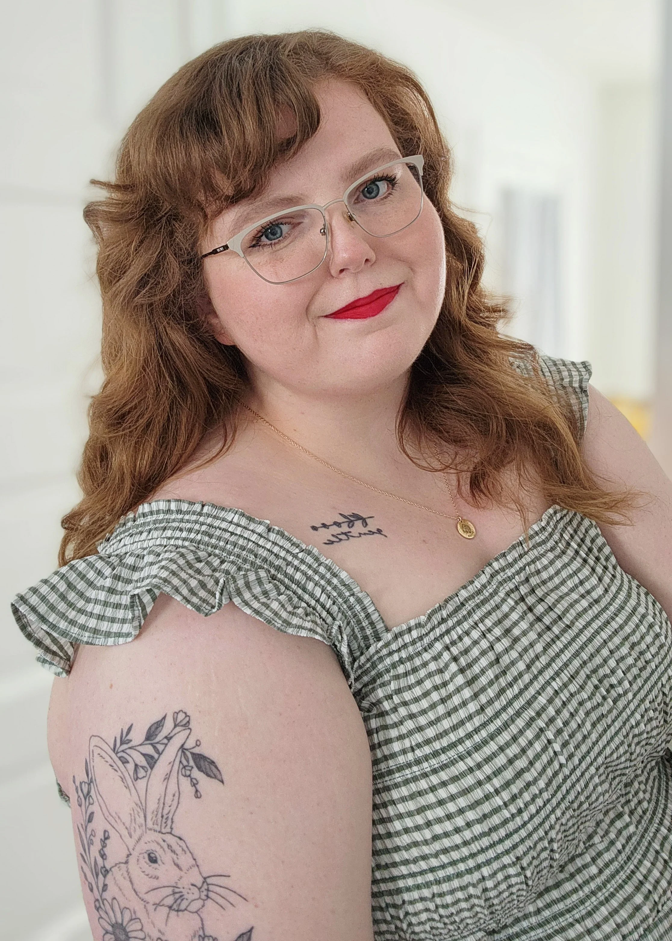 A woman with red hair, glasses, and red lipstick, wearing a checkered dress. She has tattoos of a bunny, flowers, and text on her shoulder and chest, and is smiling at the camera.
