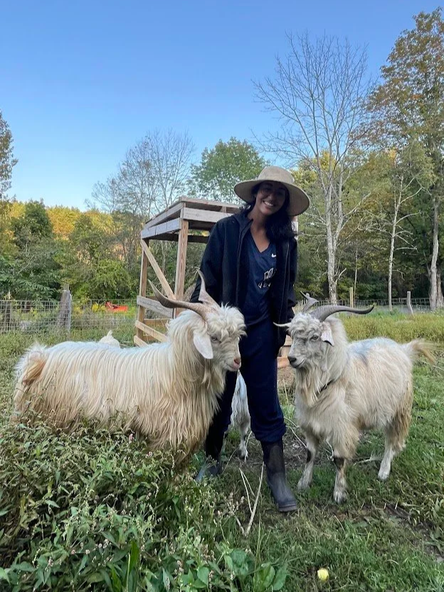 A woman wearing a wide-brimmed hat and dark clothing stands outdoors on grass, smiling and holding two large Cashmere goats with long horns.