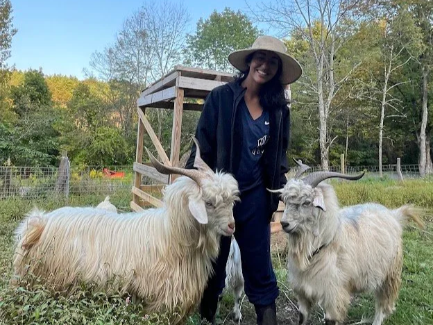 A woman wearing a wide-brimmed hat and dark clothing standing outdoors with two white cashmere goats with large horns, in a grassy area with trees and a wooden structure in the background.