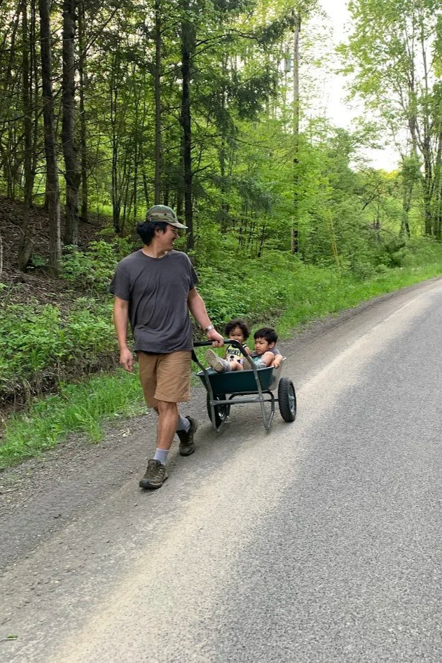 A man walking along a paved road in a wooded area, pulling a wagon with two young children inside.