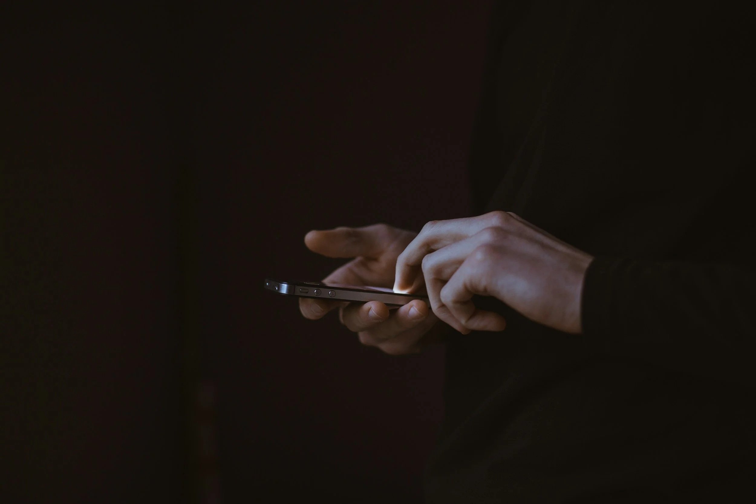 Person using a smartphone in a dark environment, only the hands and phone are visible.