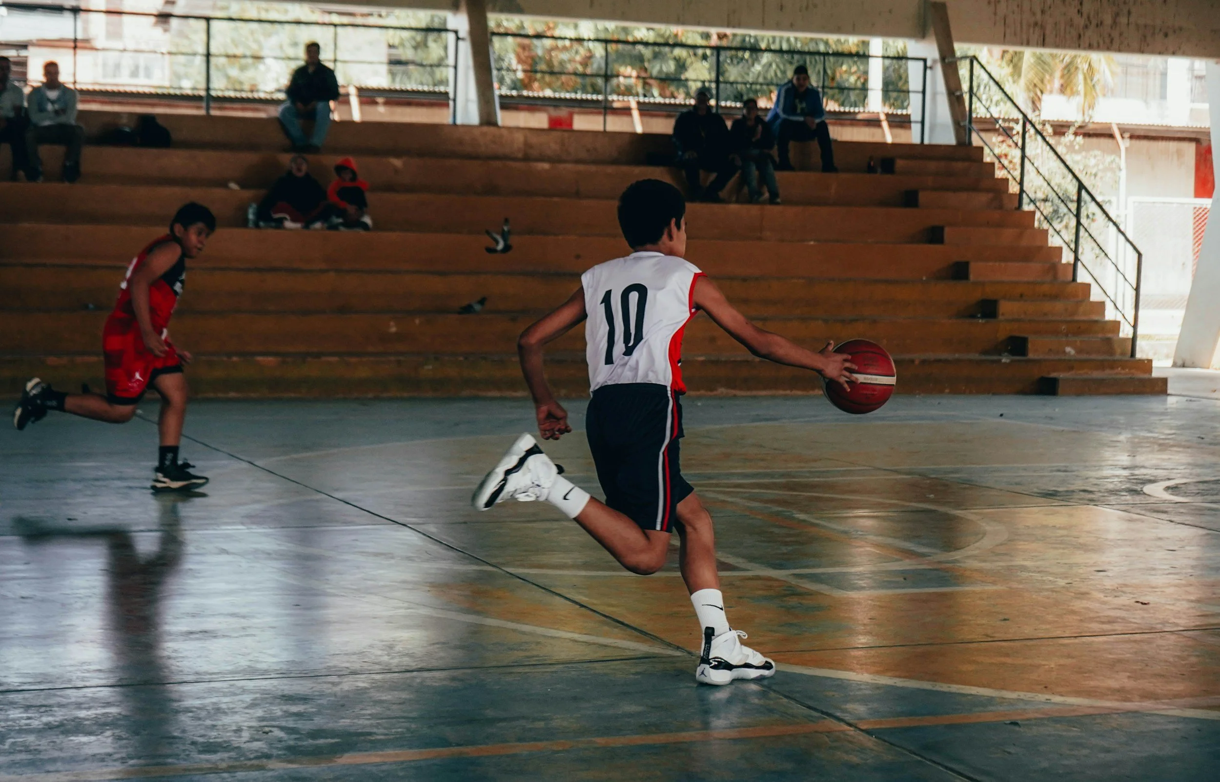 A young boy in a white and navy basketball uniform with the number 10 dribbling a basketball on an indoor court, while another boy in a red uniform runs behind him. Spectators sit on wooden bleachers in the background.