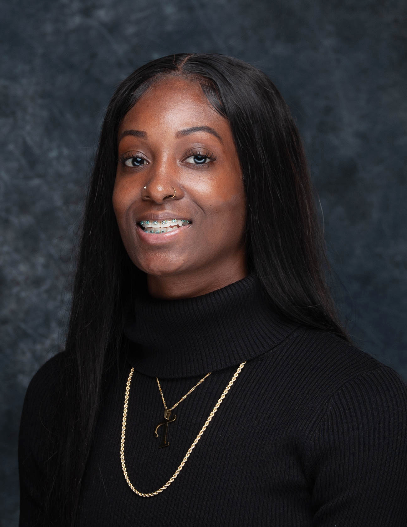 Portrait of a woman with long black hair, wearing a black turtleneck and gold jewelry, smiling lightly against a dark textured background.