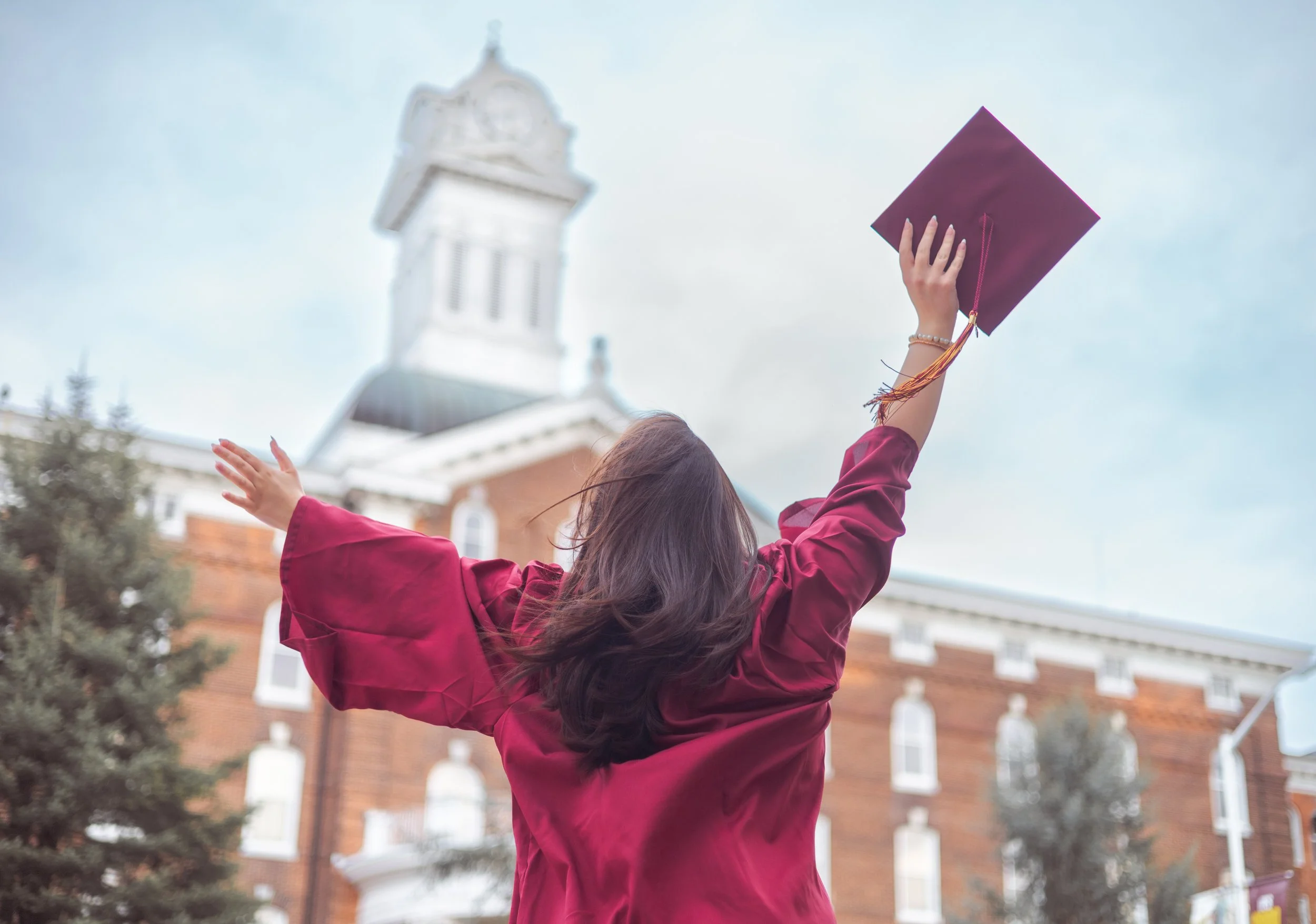 A female graduate in a maroon cap and gown holding her graduation cap up in the air, celebrating outside a brick building with white accents and a tower, with trees in the background.