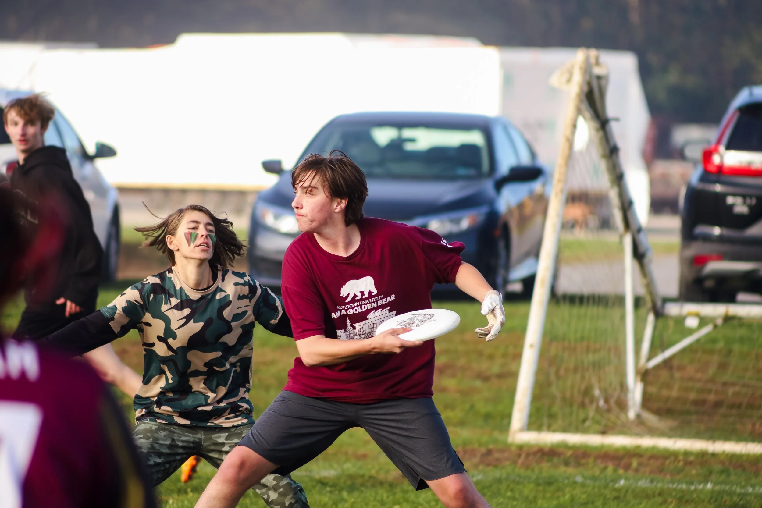 A group of young people playing ultimate frisbee on a grassy field, with cars parked in the background.