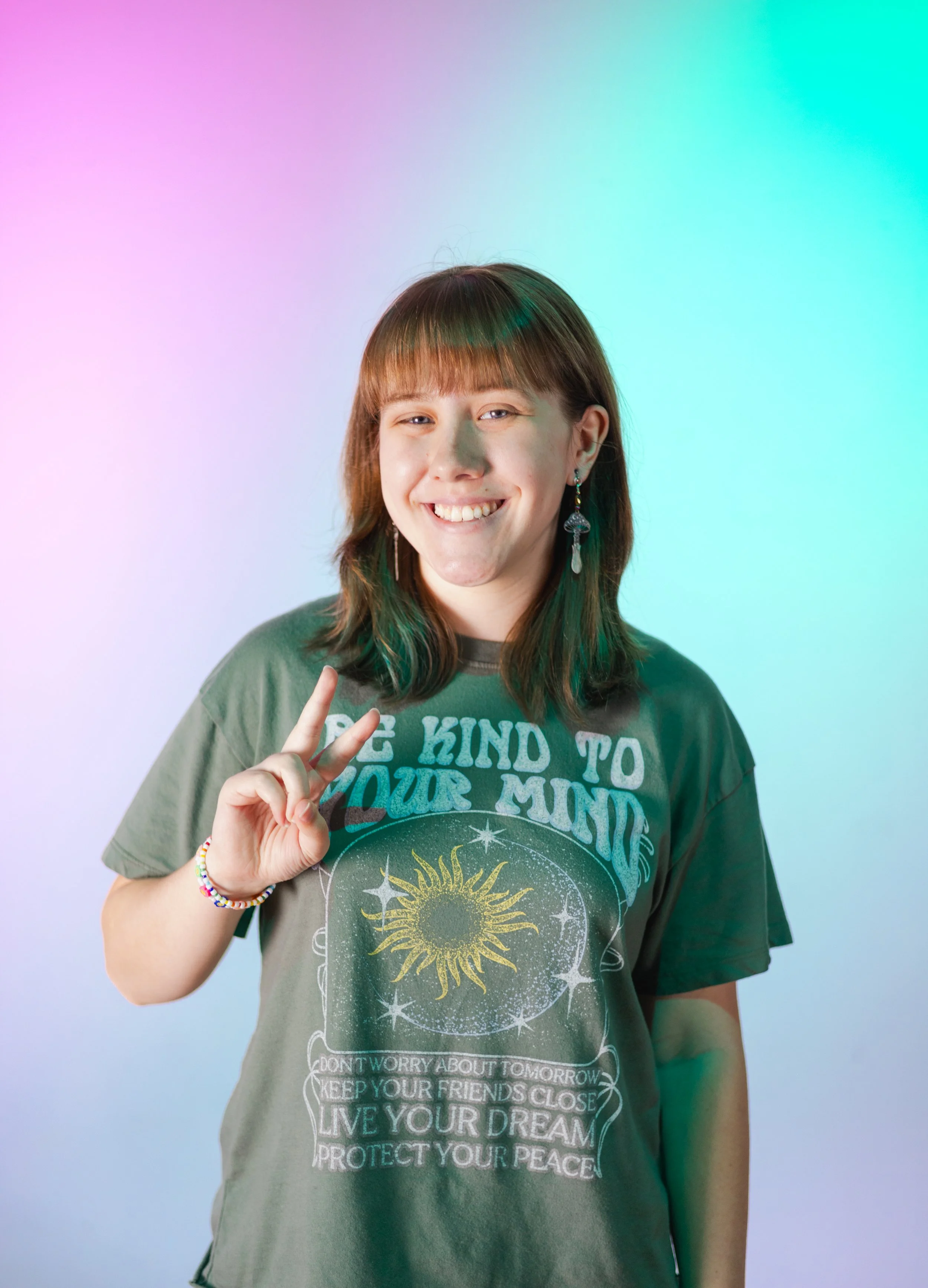 A young woman with brown hair and earrings, smiling and making a peace sign, stands against a colorful pastel background, wearing a green T-shirt with cosmic and inspirational text.