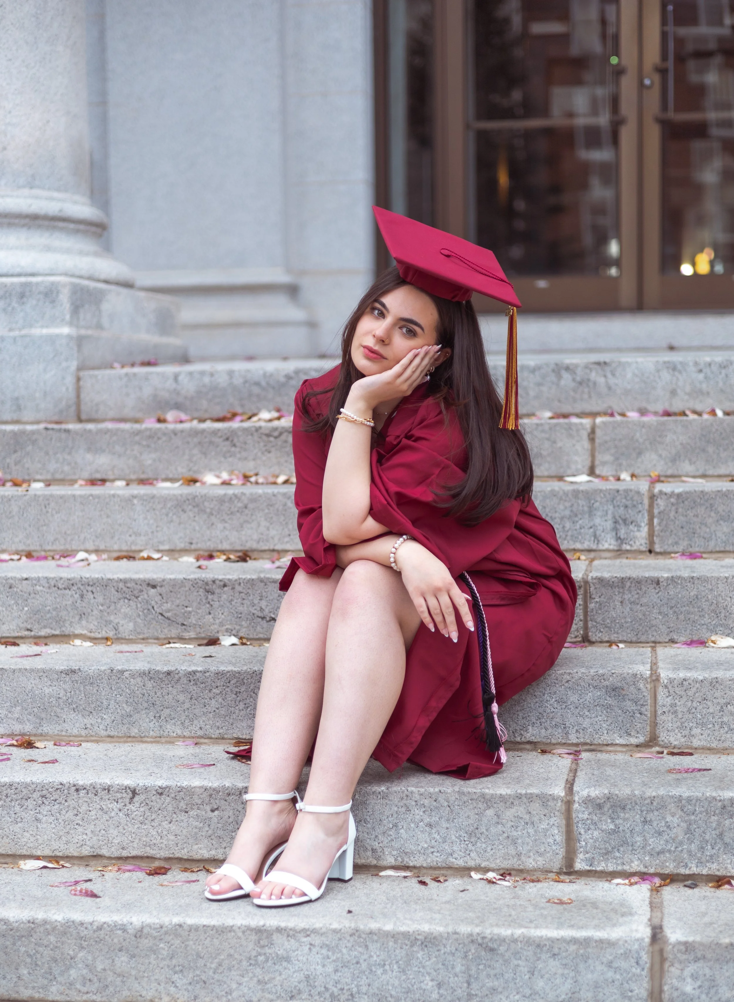 A young woman in a maroon graduation gown and cap sitting on stone steps, resting her head on her hand, with white high heels and a background of a building entrance.