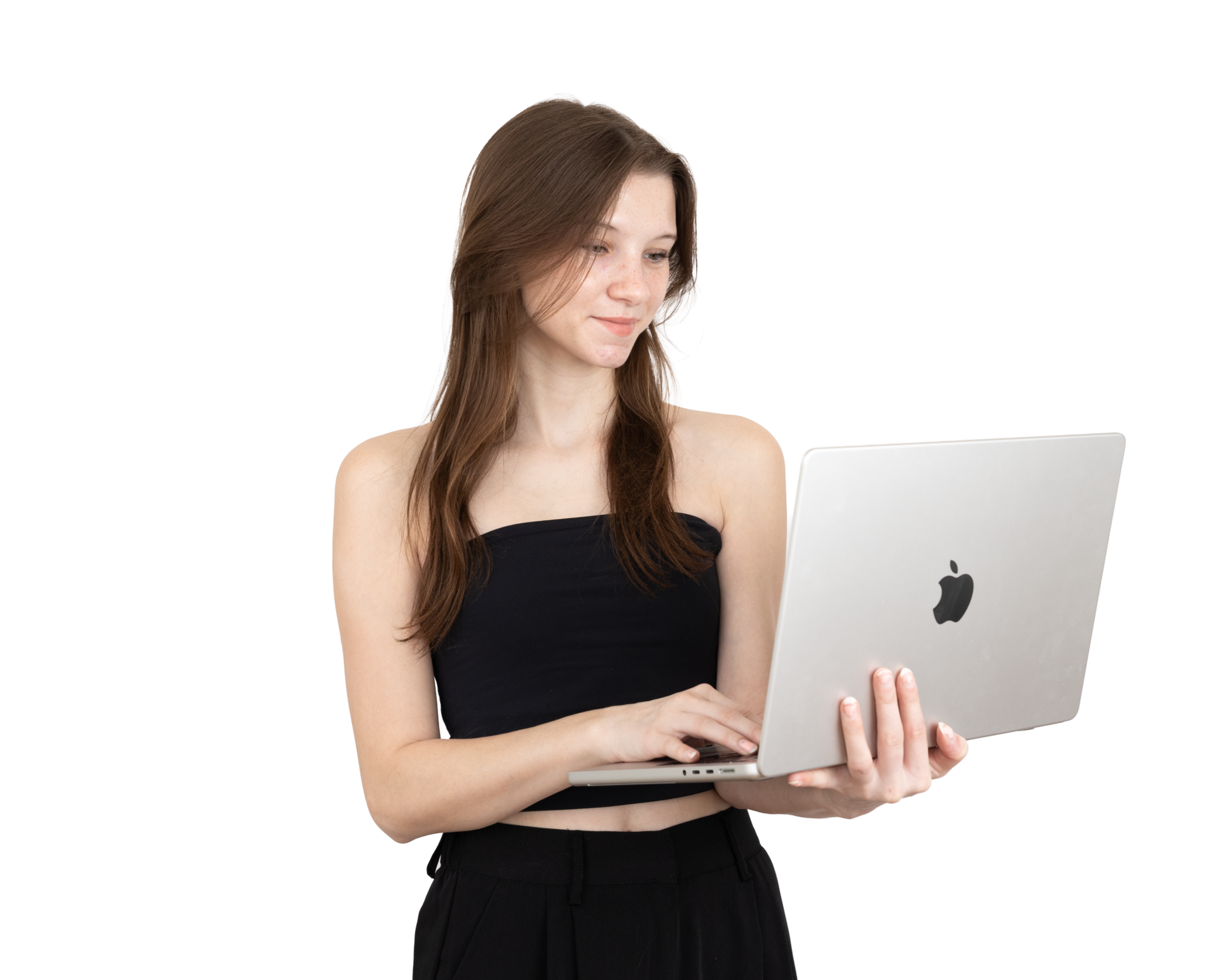 A young woman with long brown hair, wearing a black strapless top and black pants, looking at and using an open silver MacBook laptop.