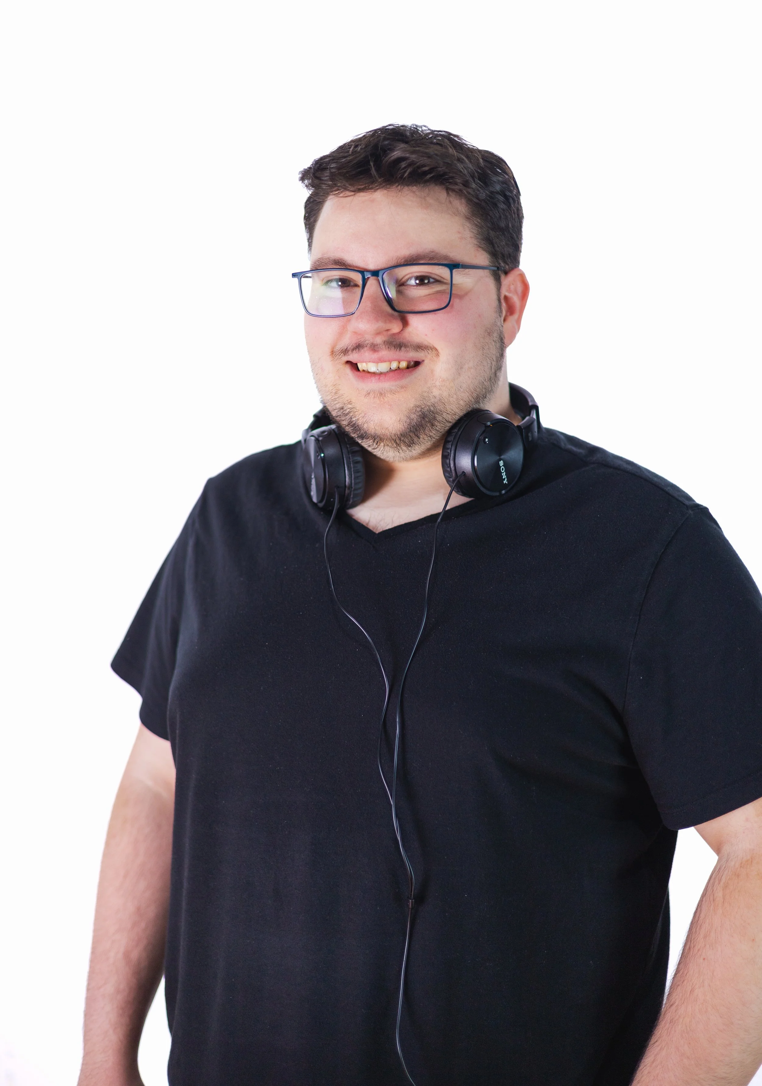 A smiling man wearing glasses and headphones around his neck, dressed in a black t-shirt, standing against a plain white background.
