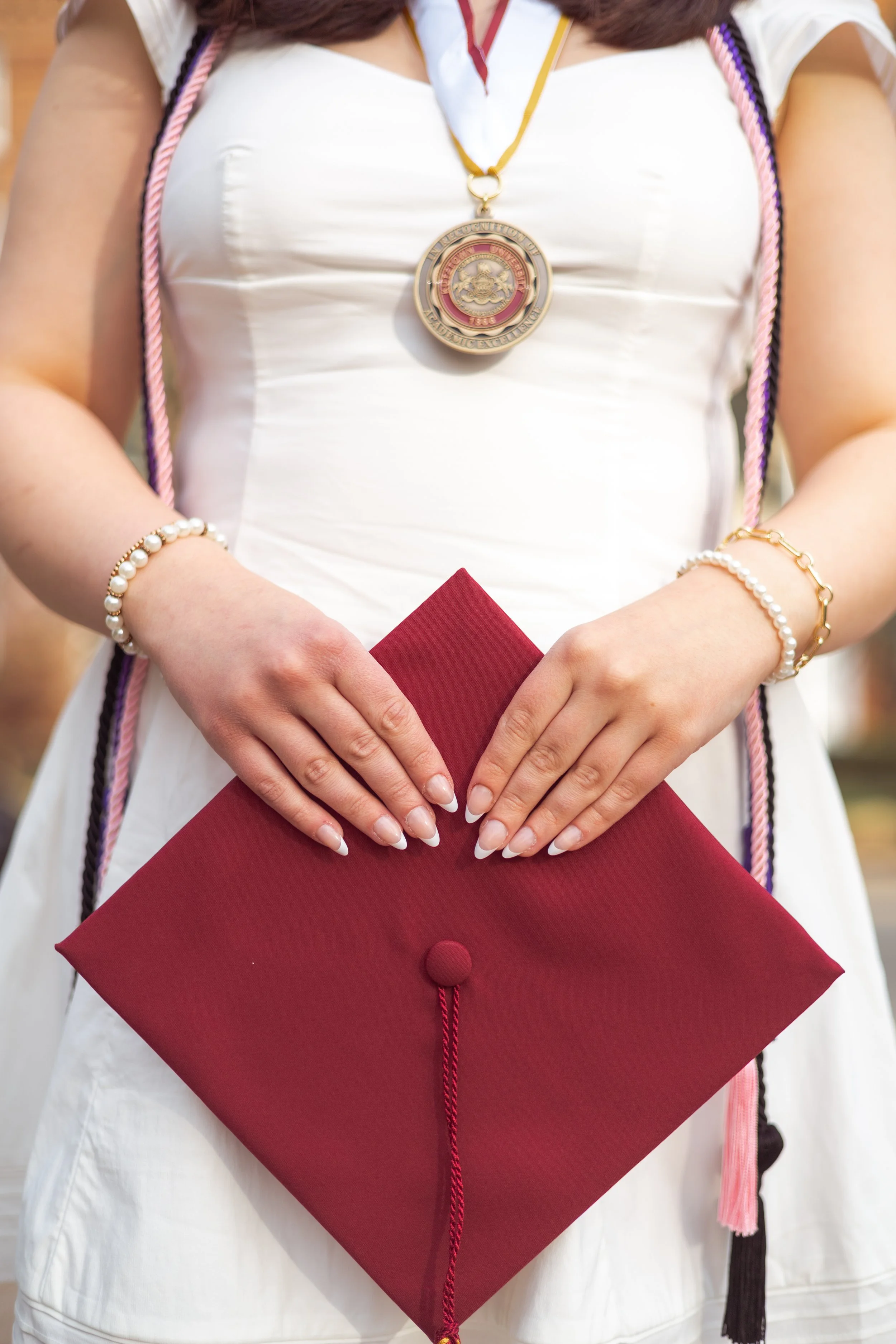 A woman in a white dress holding a red graduation cap with both hands. She is wearing a medallion around her neck, pearl bracelets, and her nails are manicured.