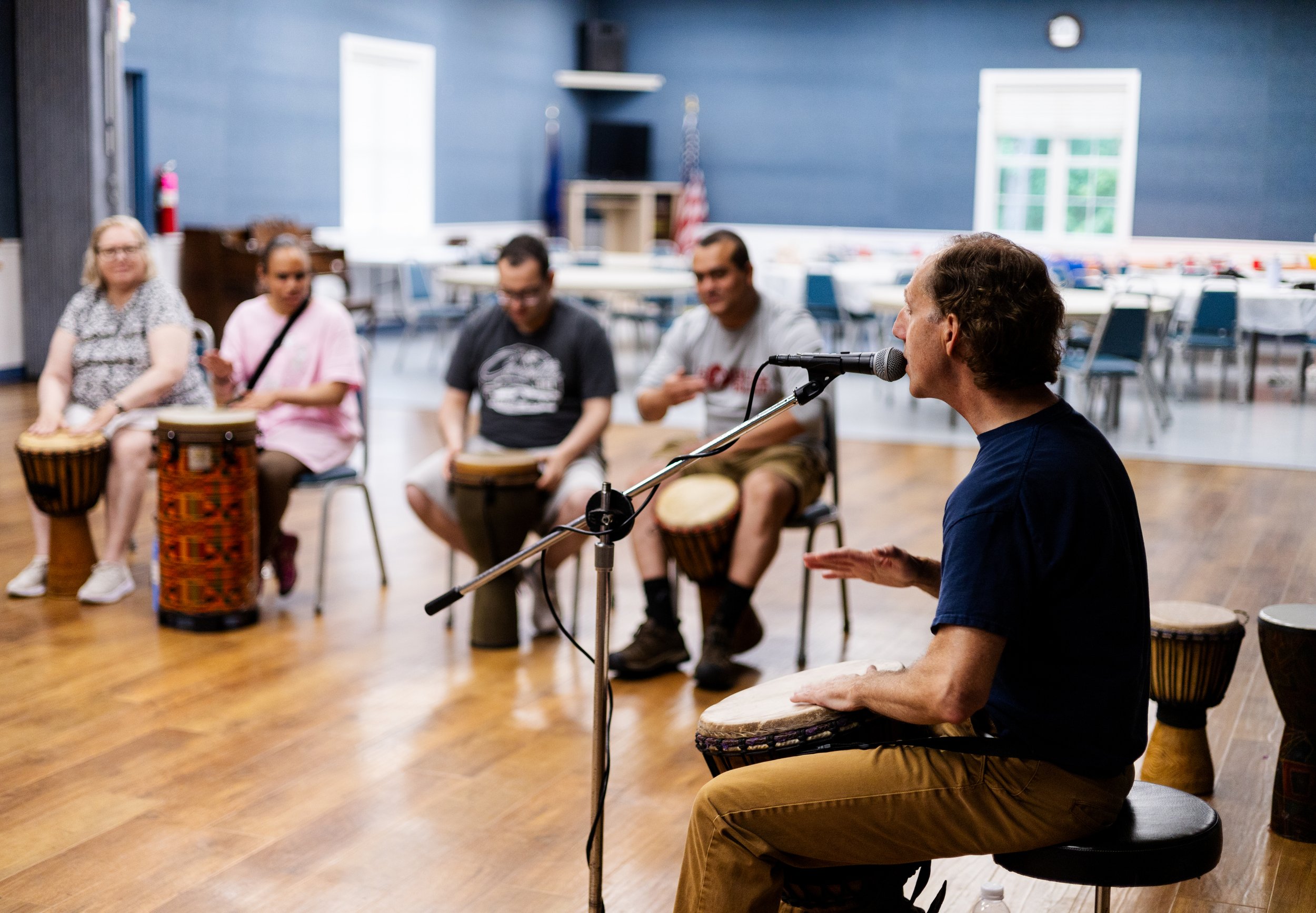 A group of people sitting on chairs in a room, participating in a drumming workshop led by a man at a microphone, with drums in the background and a blue wall.
