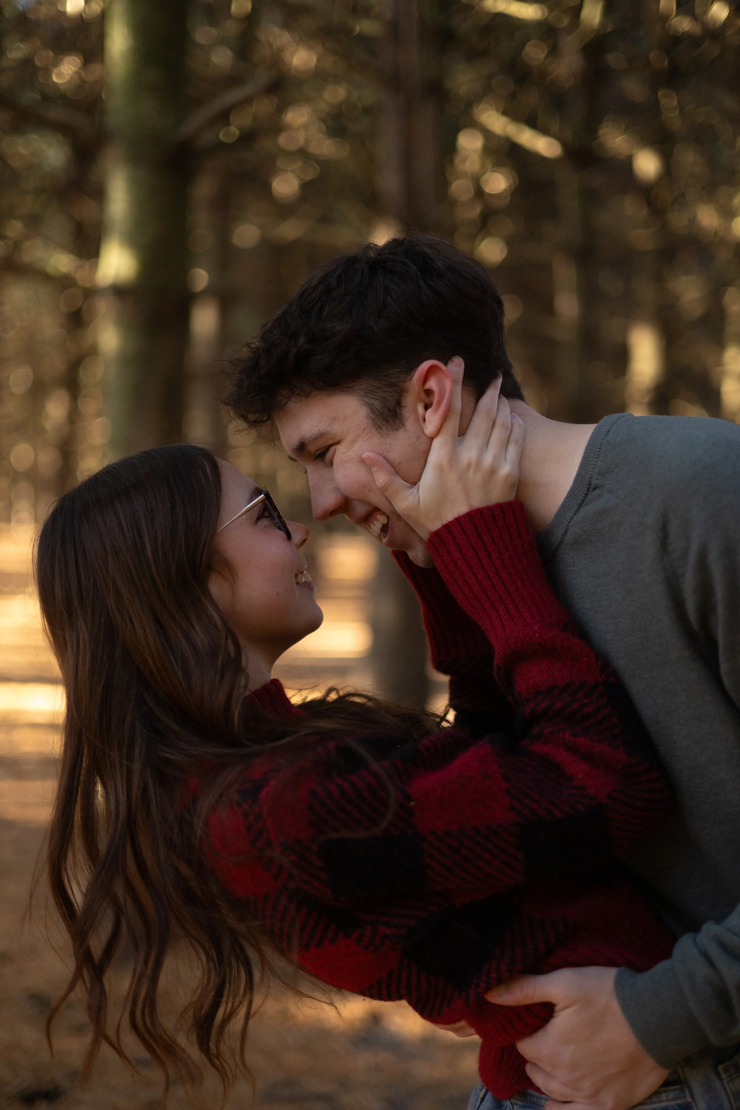 A couple is embracing and smiling at each other in an outdoor forest setting during the daytime.