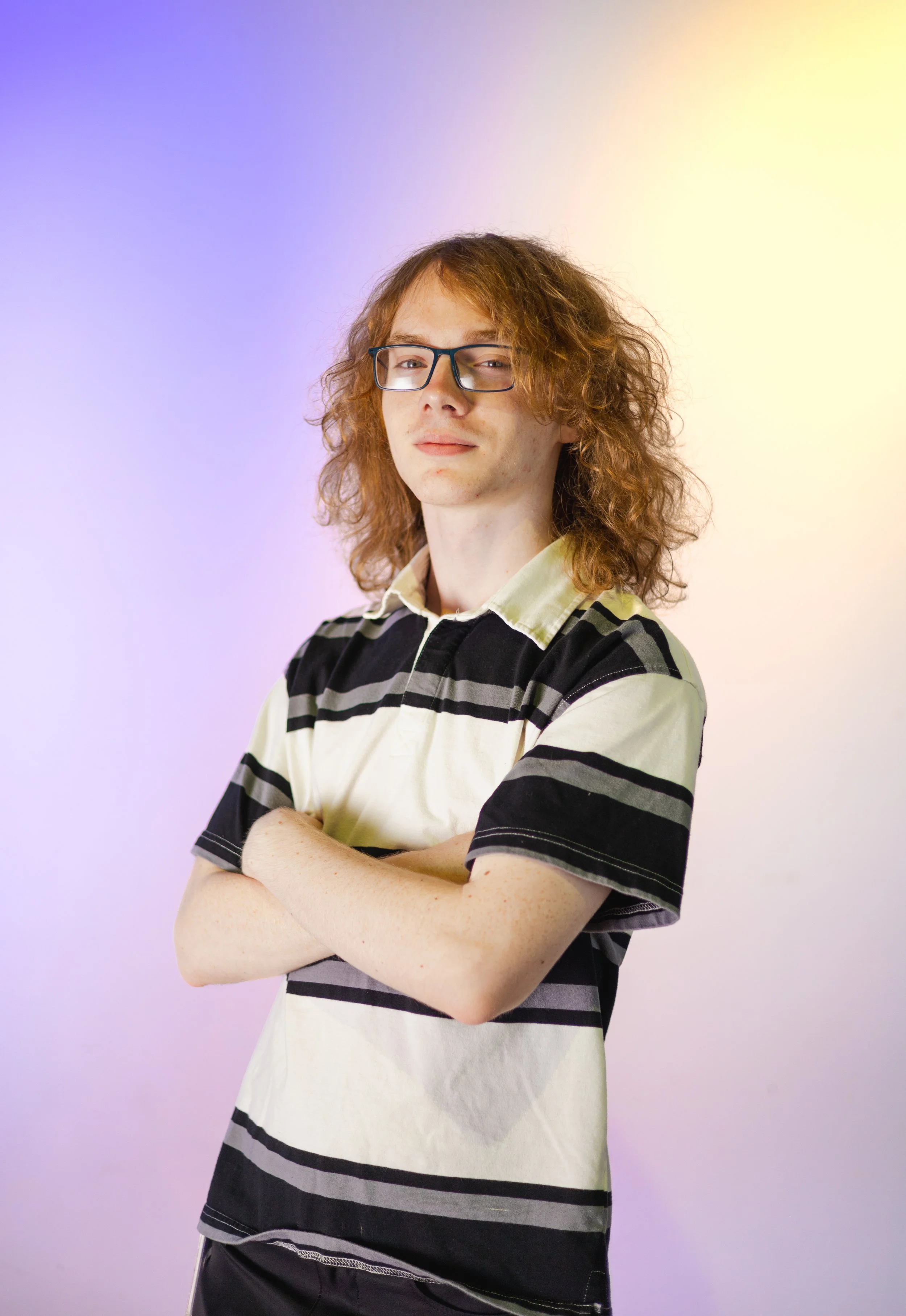 A young man with curly red hair and glasses standing with arms crossed against a softly colored background in shades of purple, pink, and yellow.