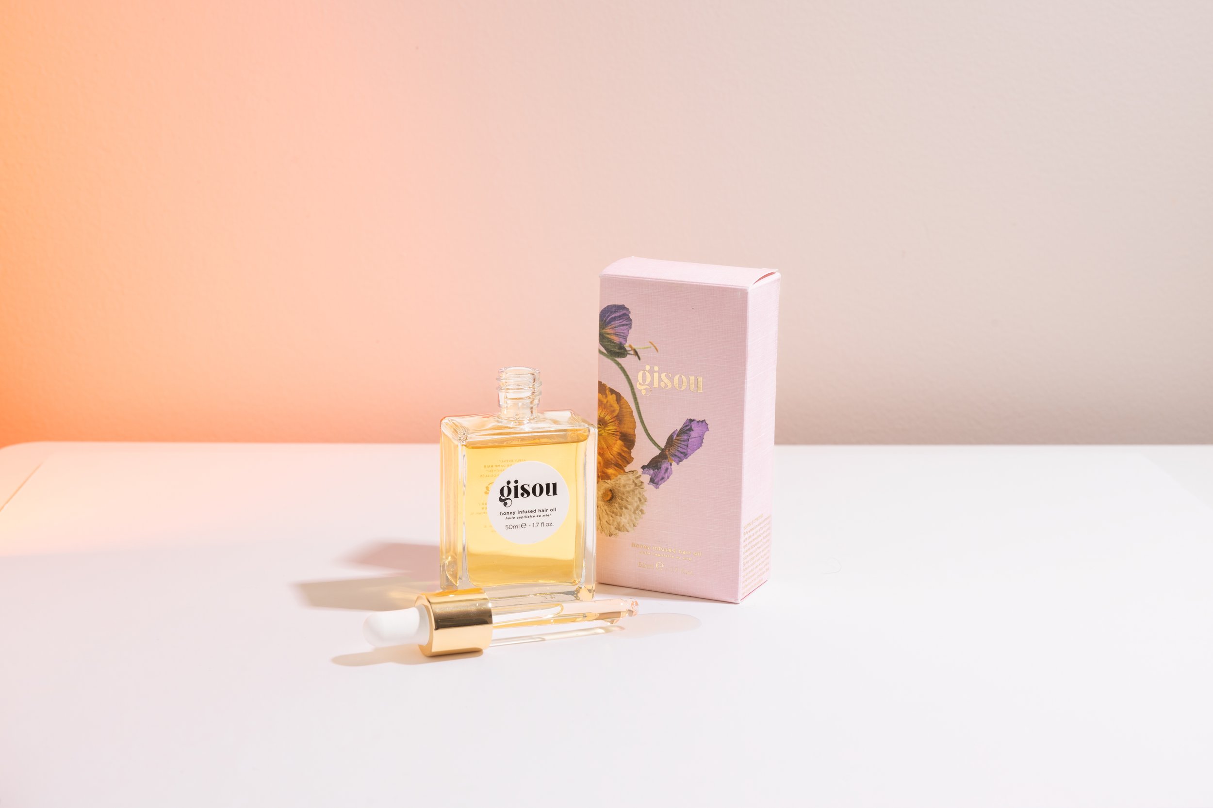 A bottle of Gisou honey-infused hair oil with its box and a dropper on a white surface, against a softly lit background.