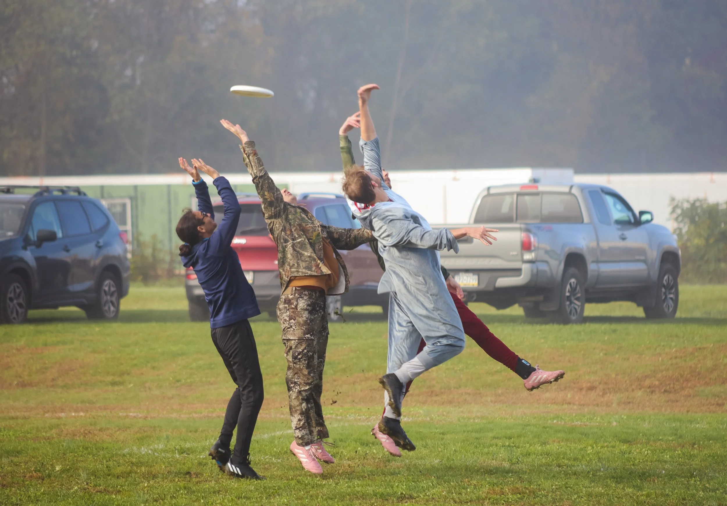 Four people jumping in the air with their hands raised, trying to catch a frisbee. They are standing on grass with parked cars in the background.
