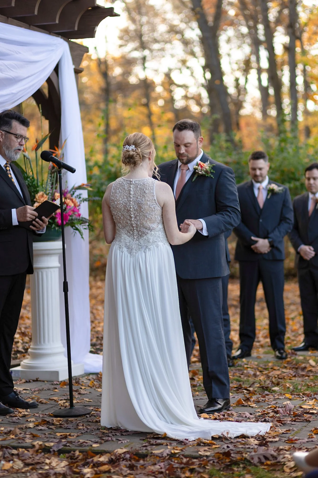 A couple is exchanging wedding vows outdoors during a fall wedding ceremony. The bride is wearing a sleeveless, beaded gown, and the groom is in a dark suit with a flower boutonniere. Four men are standing in the background, dressed in suits with bou